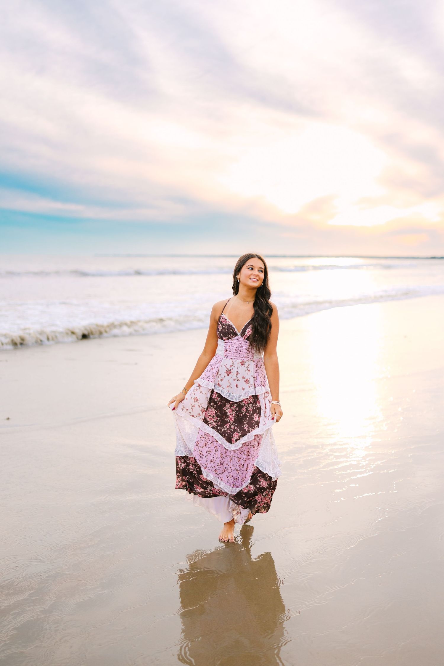 Young woman in a floral maxi dress walking along a reflective beach at a vibrant sunset.