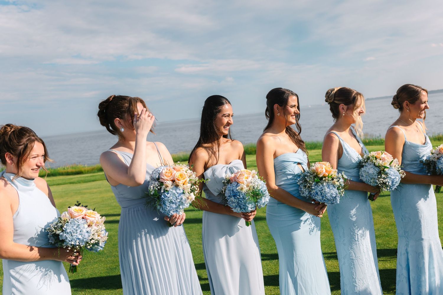 Bridesmaids in light blue dresses laughing and holding blue and white floral bouquets outdoors.
