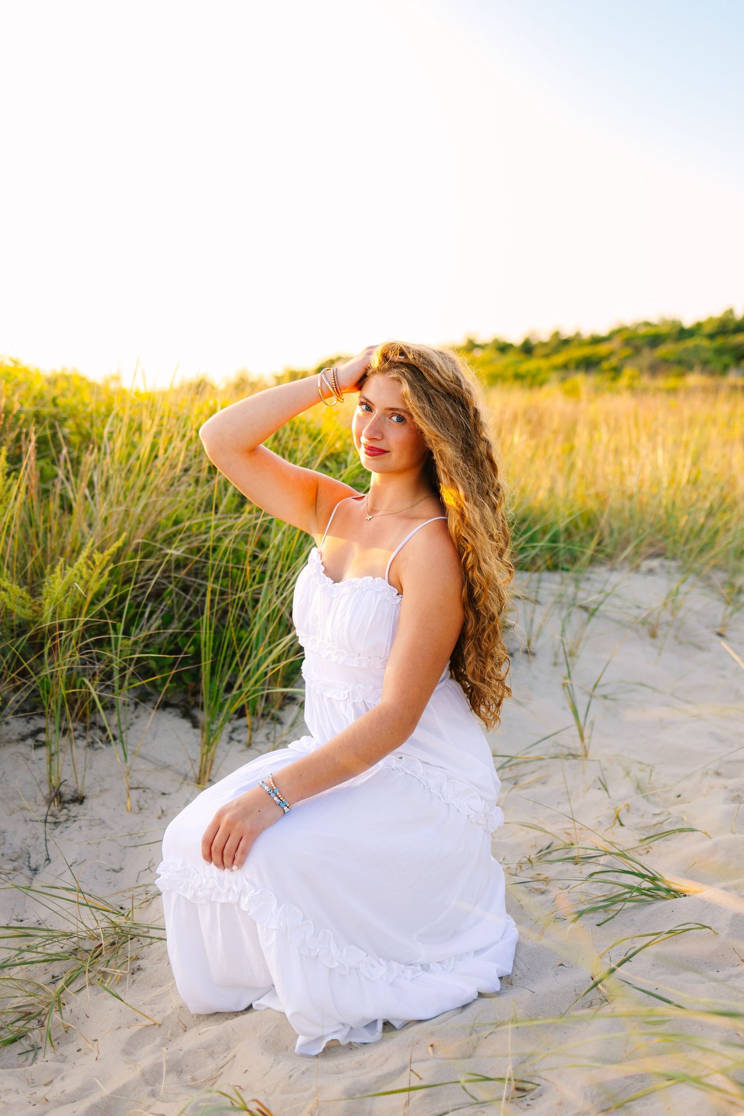 Young woman in white dress sitting on sandy beach dunes at golden hour.