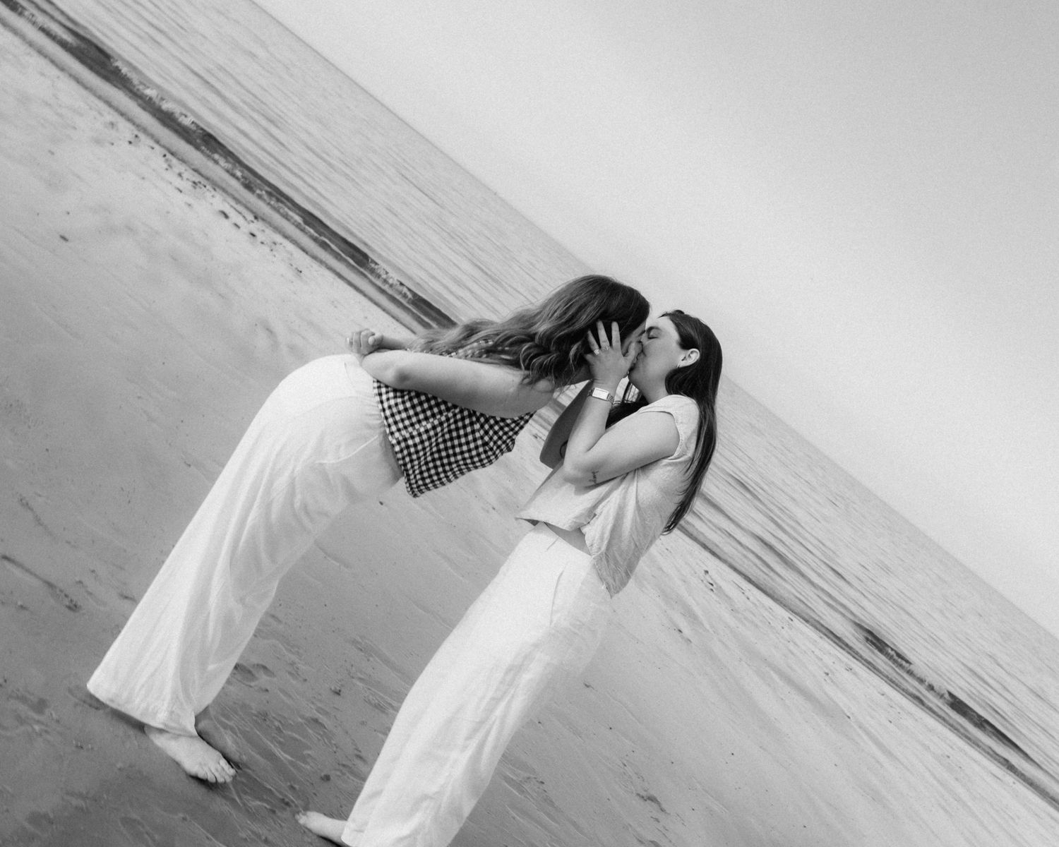 Black and white photo of two women sharing a romantic kiss on a beach shoreline.