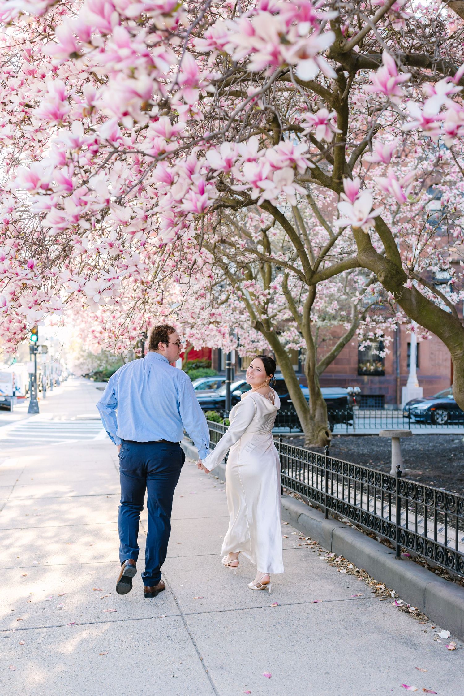A couple walks hand in hand beneath stunning pink magnolia blossoms on a charming city sidewalk.