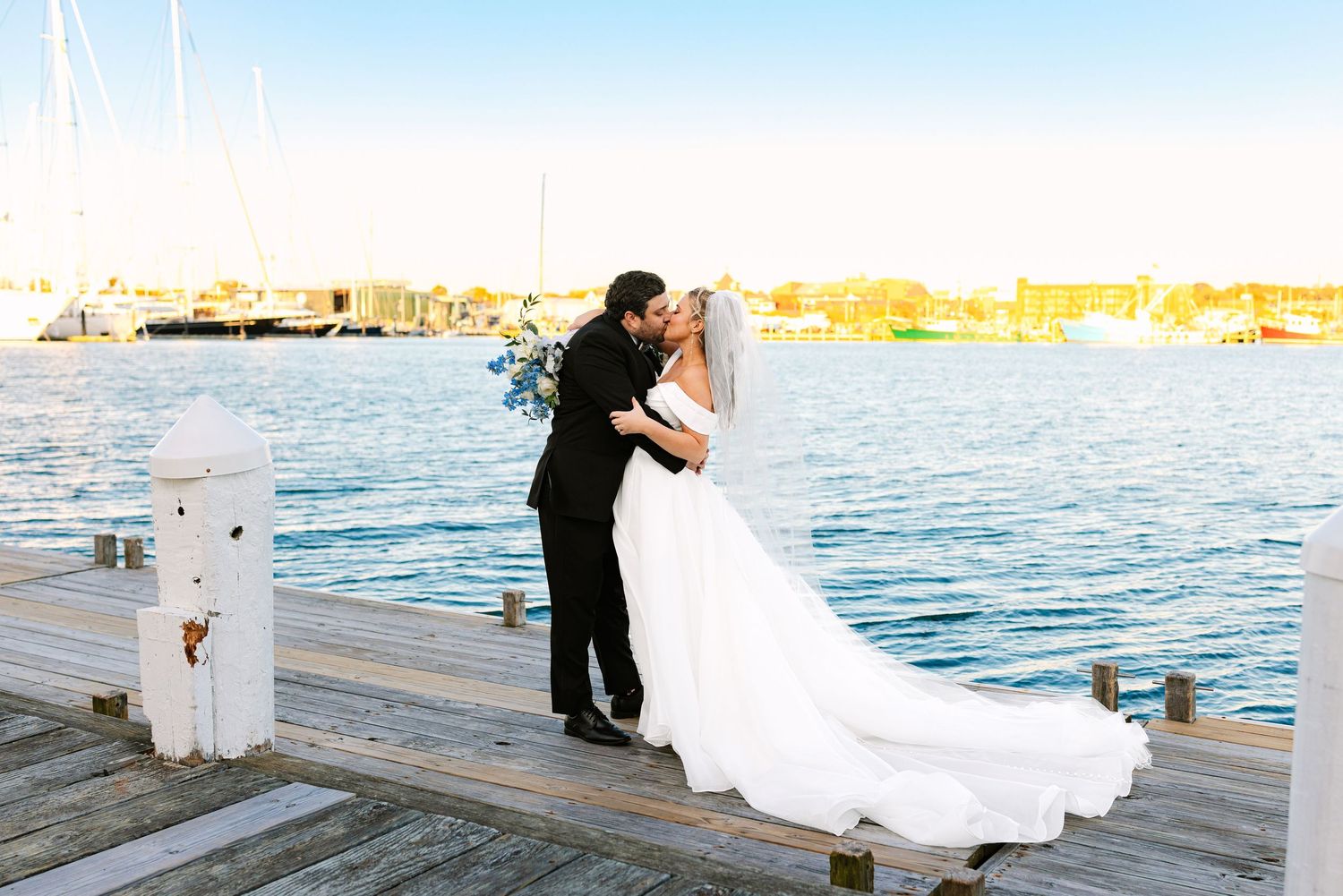 A couple in formal wedding attire dances on a wooden pier by the ocean during sunset.