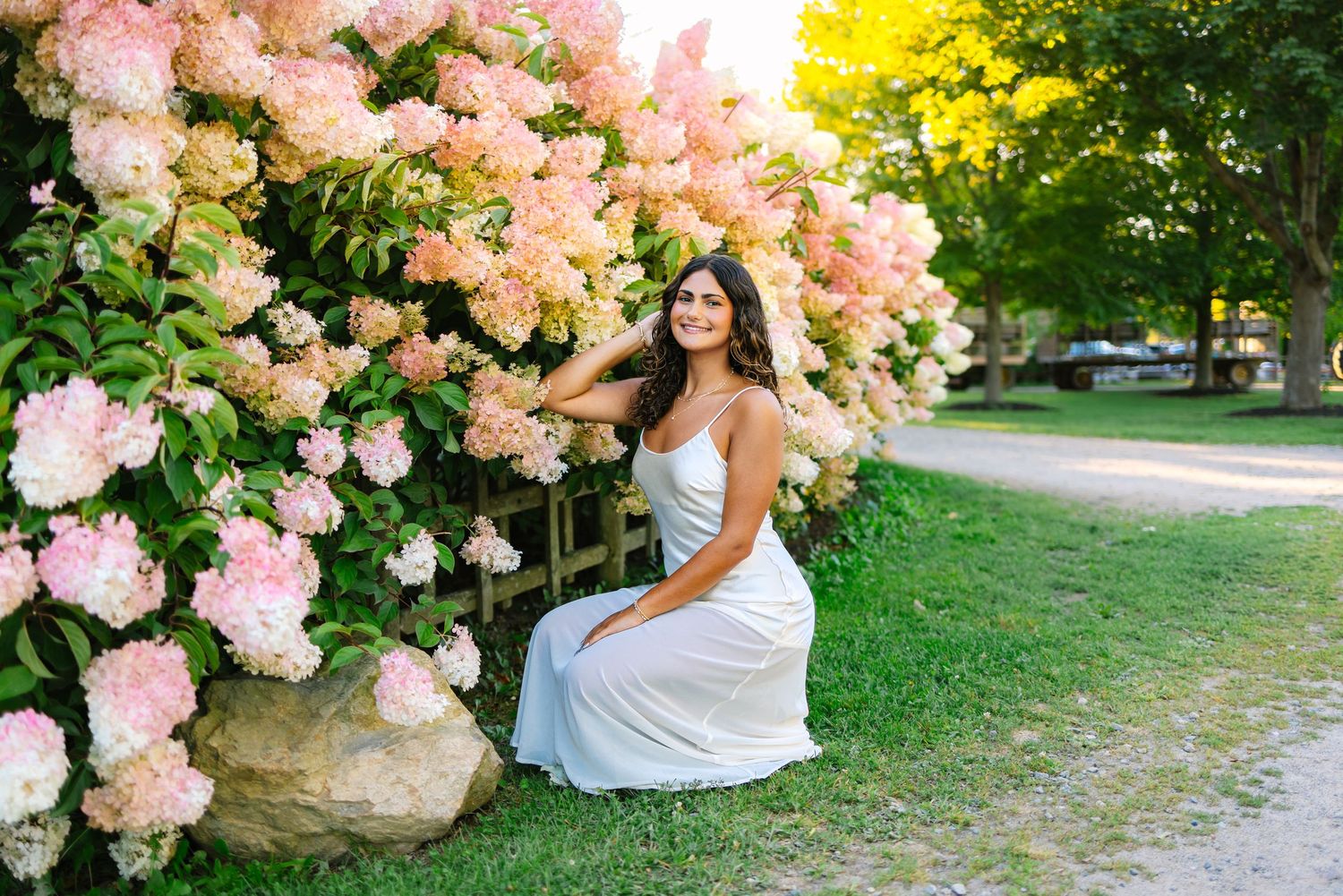 Young woman in white dress sitting on stone beside vibrant pink and white hydrangea garden bushes.