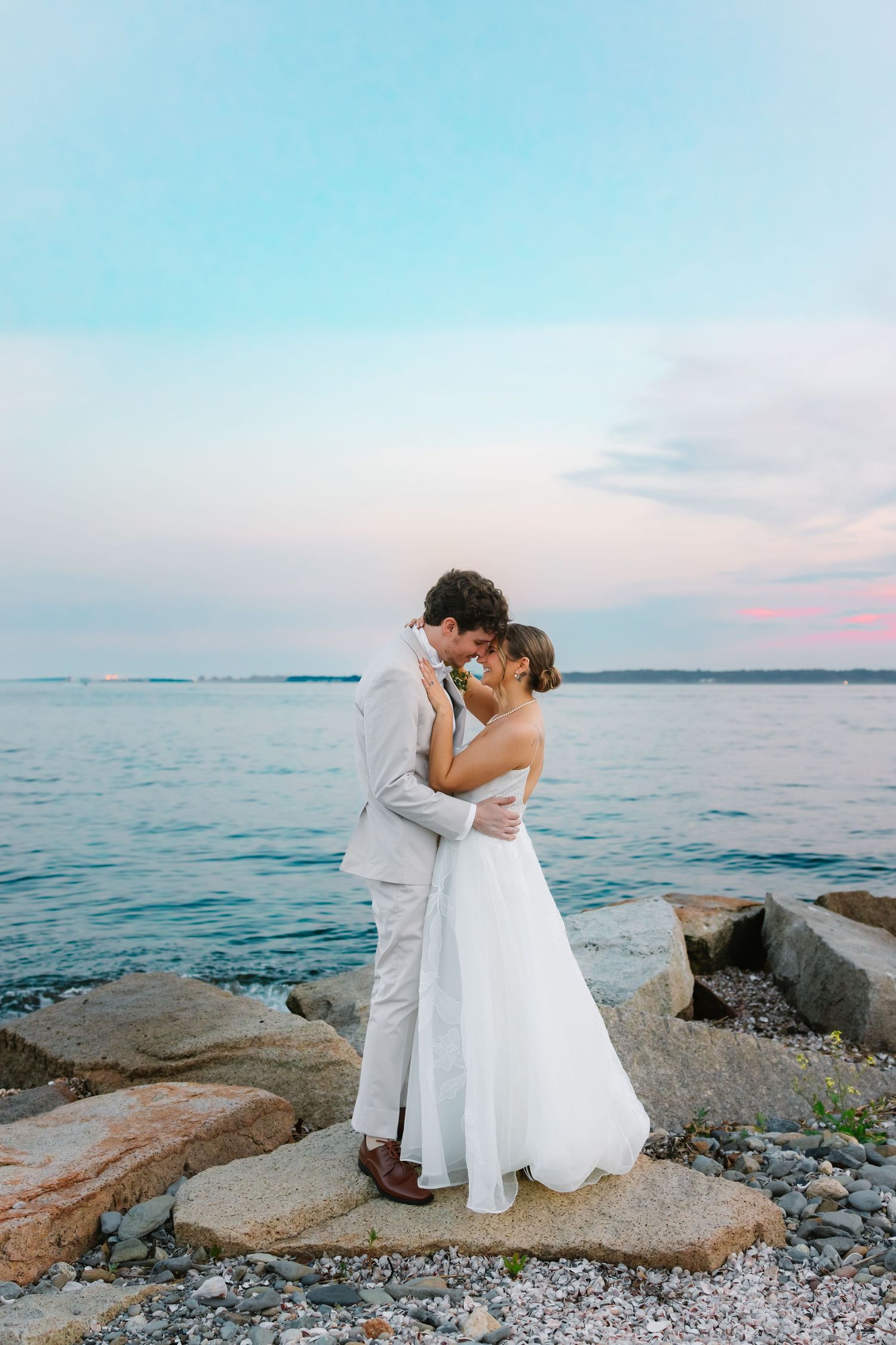 Newlyweds embracing on rocky shoreline at sunset, bride in white gown and groom in gray suit.