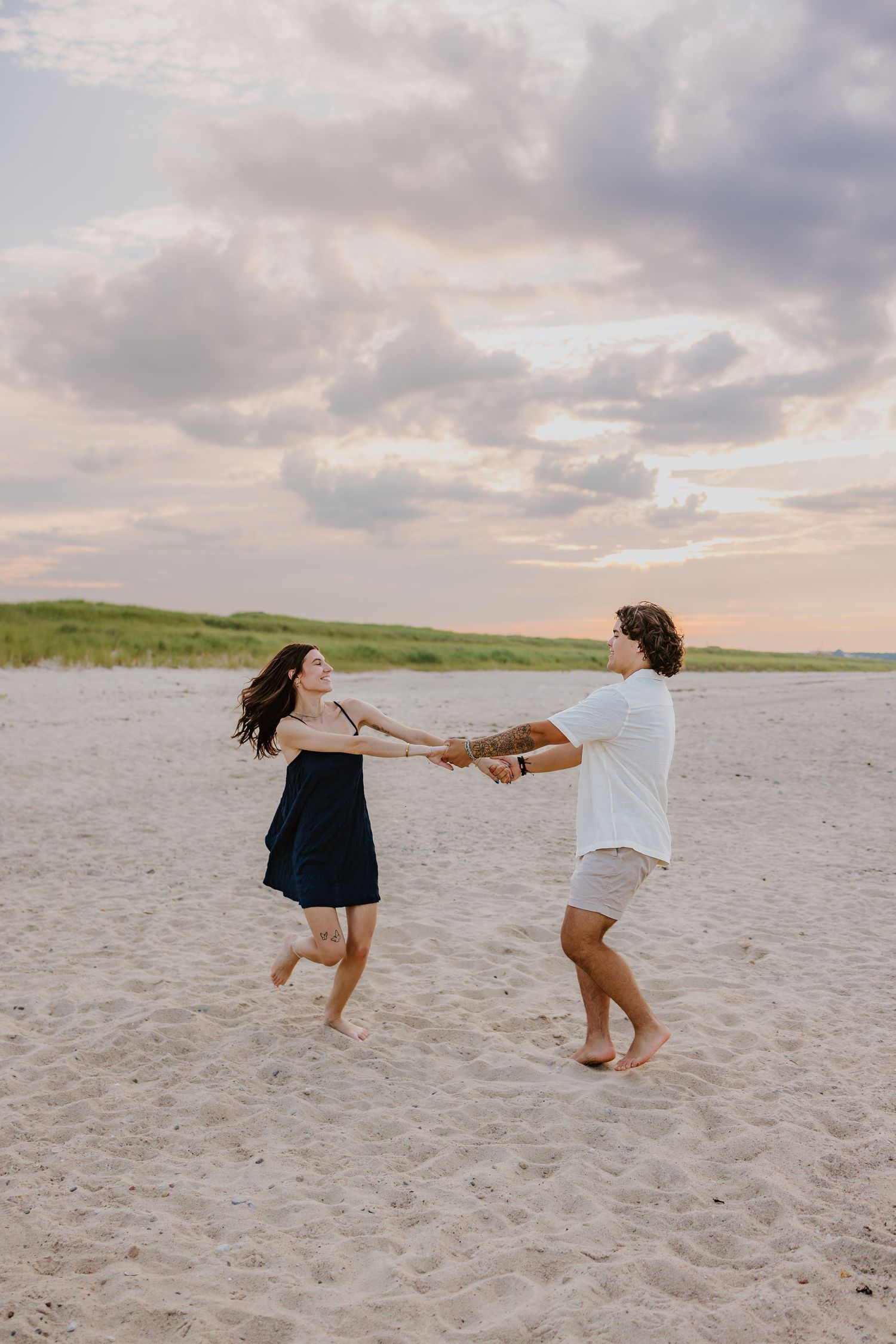 Joyful couple dancing and spinning together on sandy beach at sunset with dramatic cloudy sky.