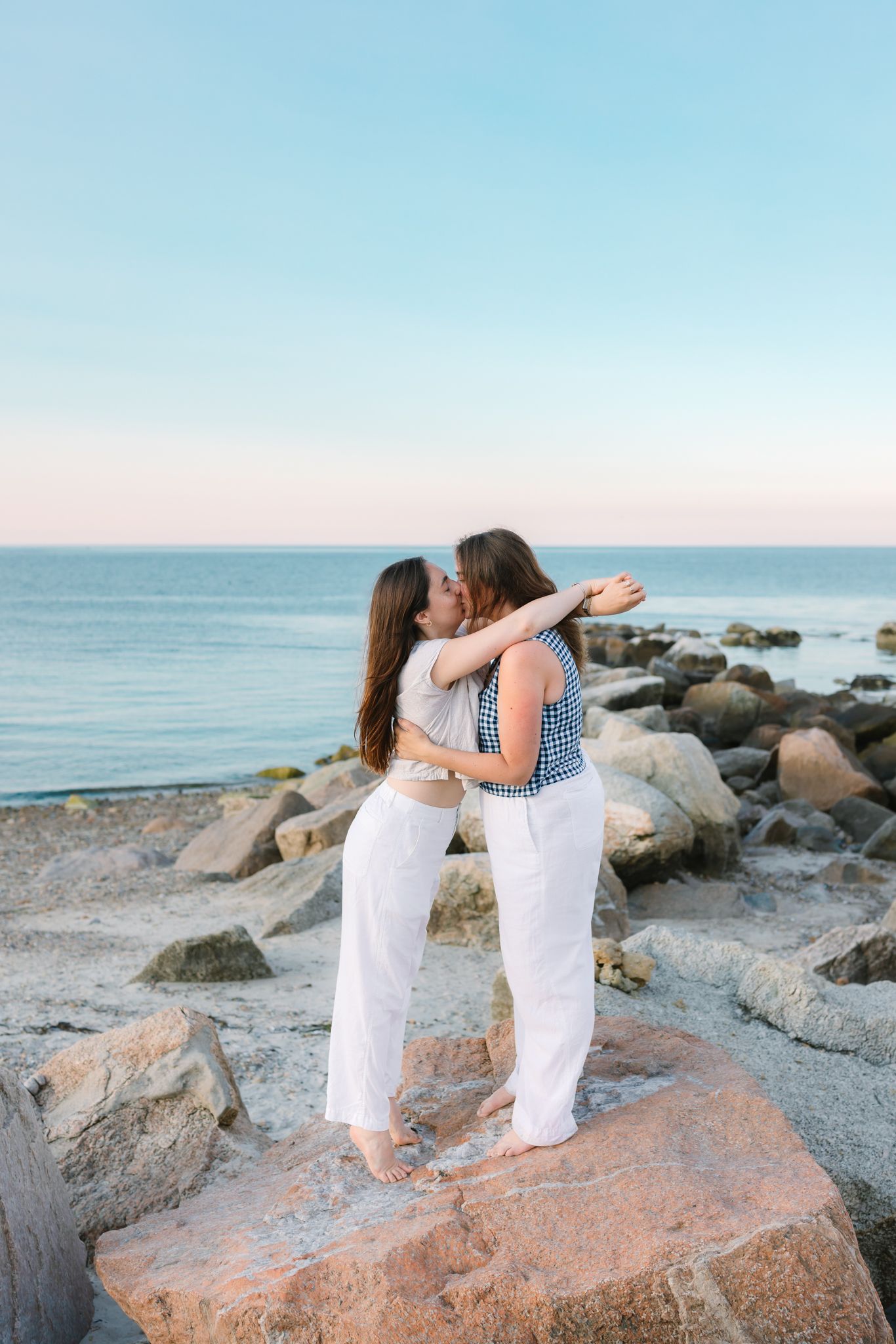 Two engaged women sharing a romantic kiss while standing on rocks by the ocean at sunset.