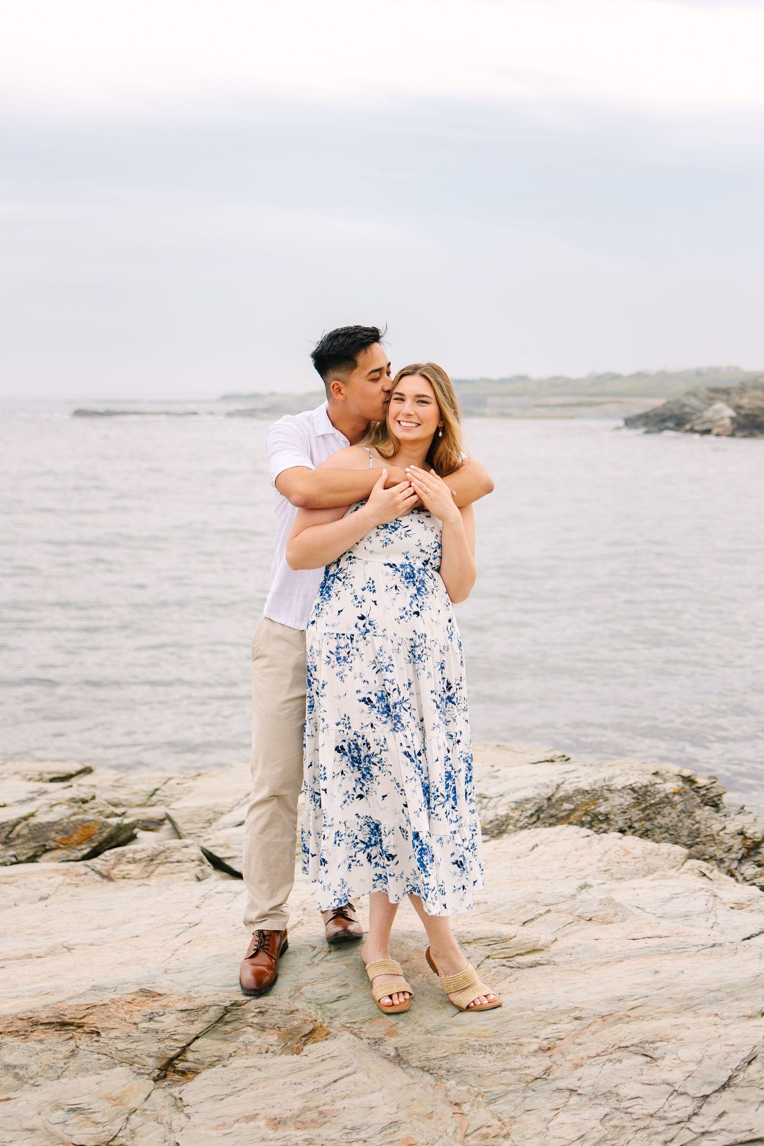 Couple embracing on rocky New England coastline for engagement photos, man kissing woman with ocean backdrop in Newport Rhode Island