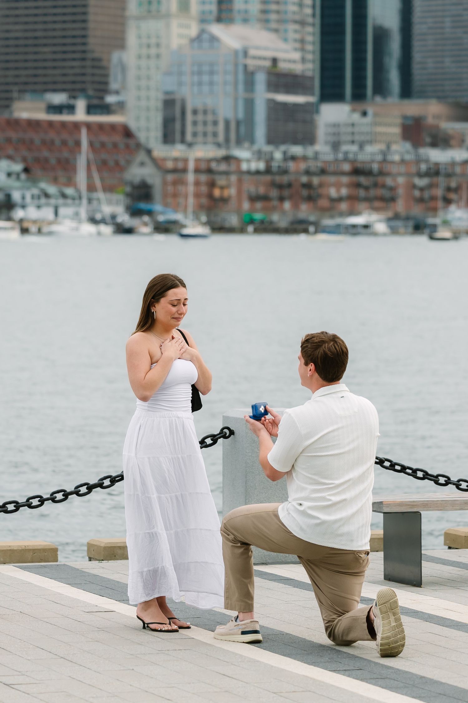 Man proposing on one knee with a ring box to a woman on a waterfront pier.