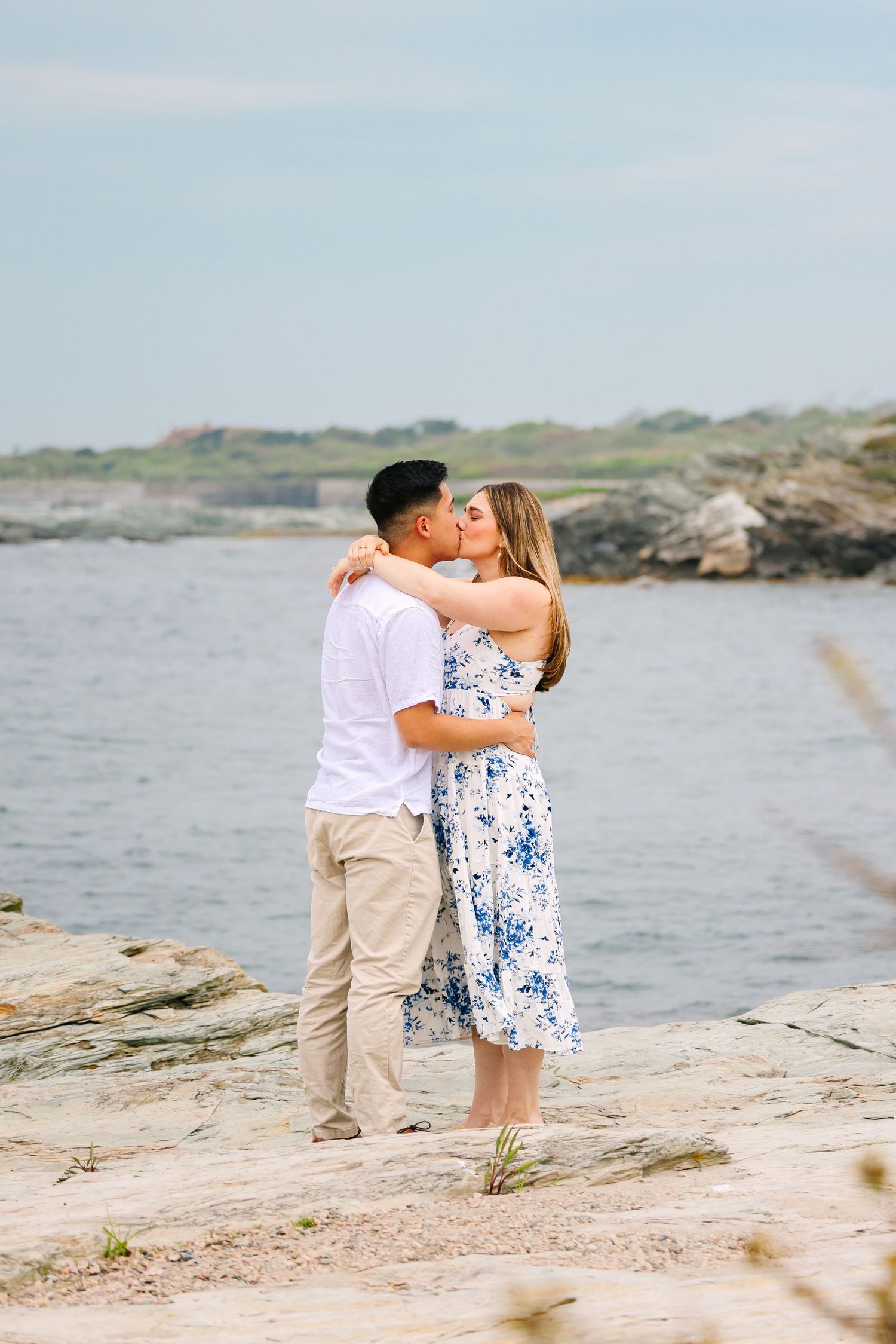 Newly engaged couple kissing and embracing on rocky coastal shoreline in Newport RI, man in white shirt and woman in blue floral dress.
