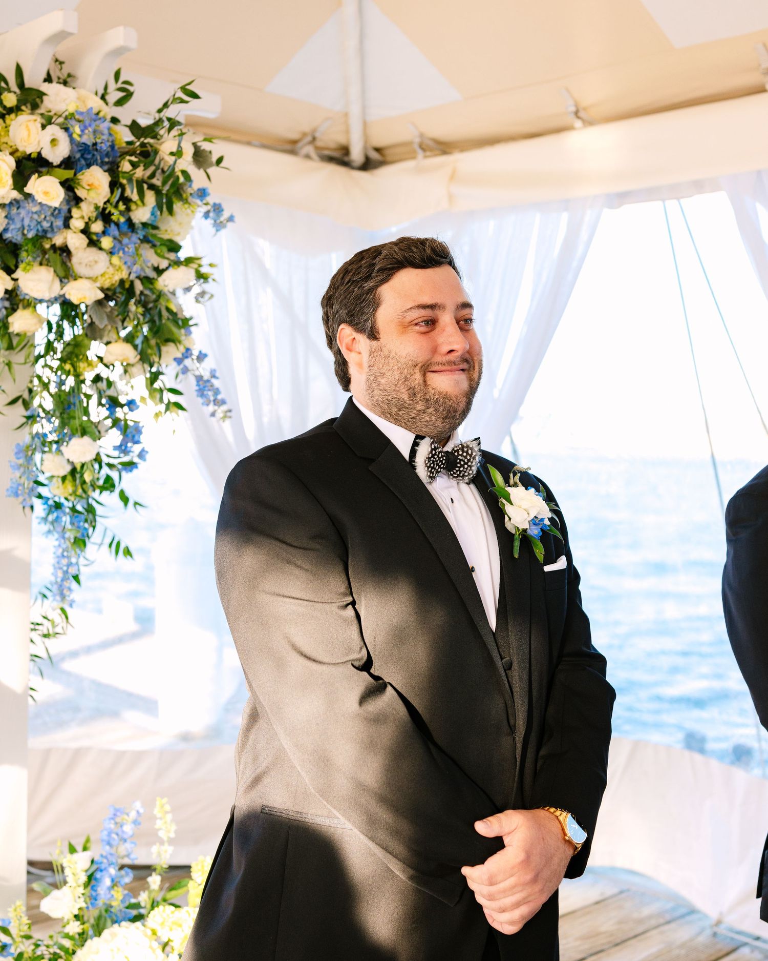 A person in a black suit laughs while standing in a white tent decorated with floral arrangements and greenery.