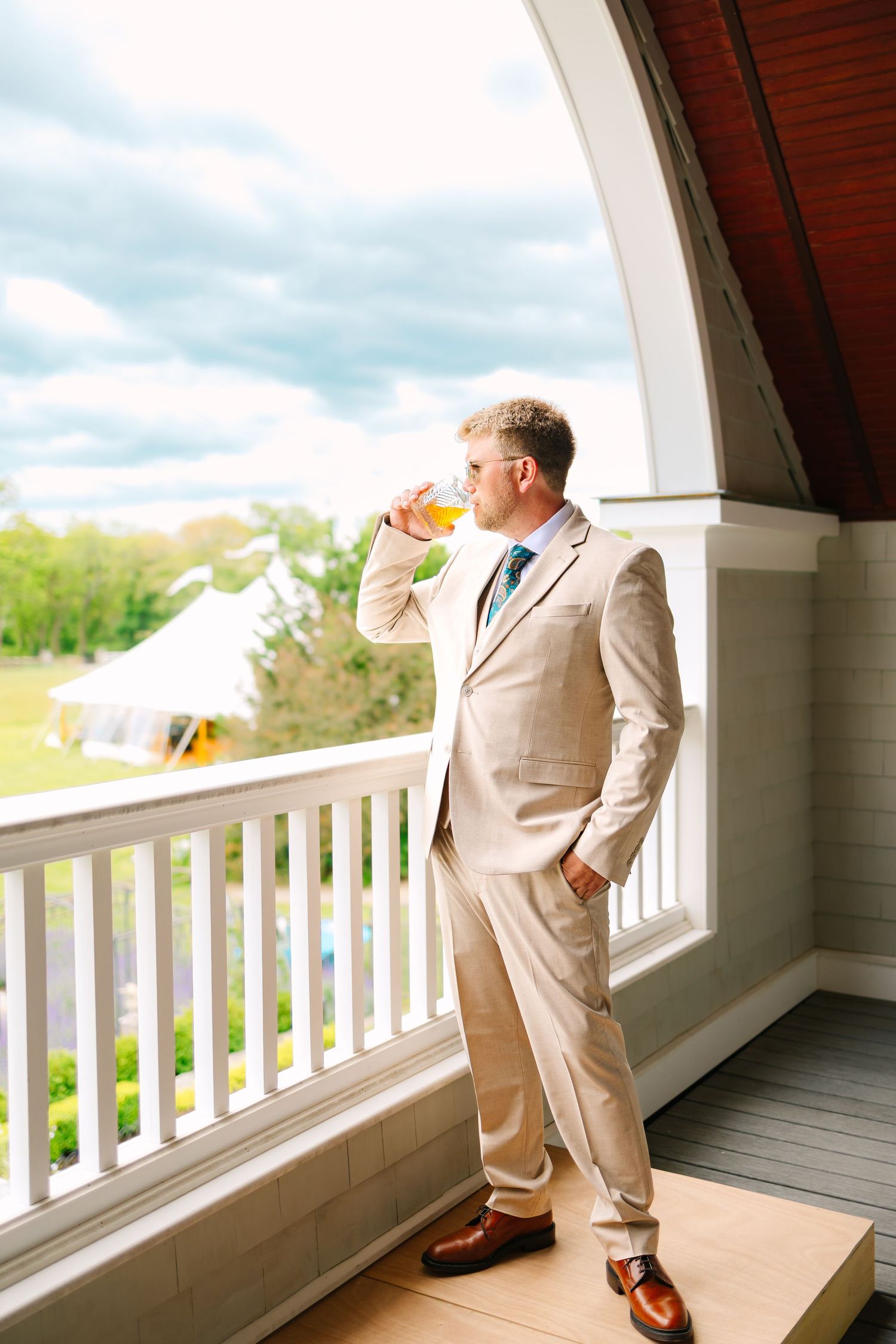 A groom in a beige suit drinks from a glass while standing on an elegant covered porch with white railings for backyard wedding in CT