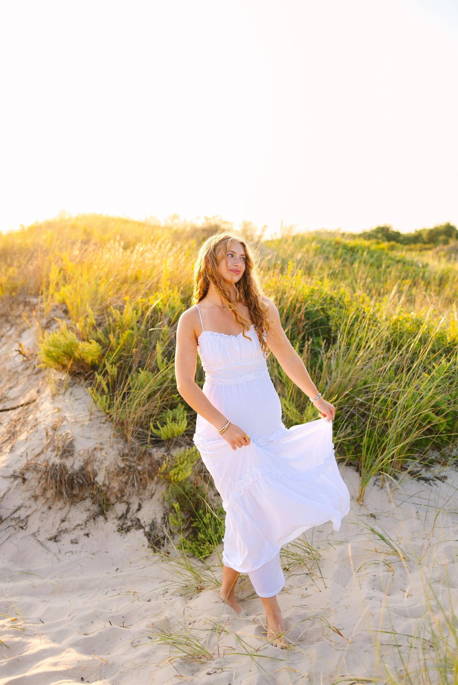 Young woman in a flowing white maxi dress walking barefoot through golden sunset sand dunes with tall grass.