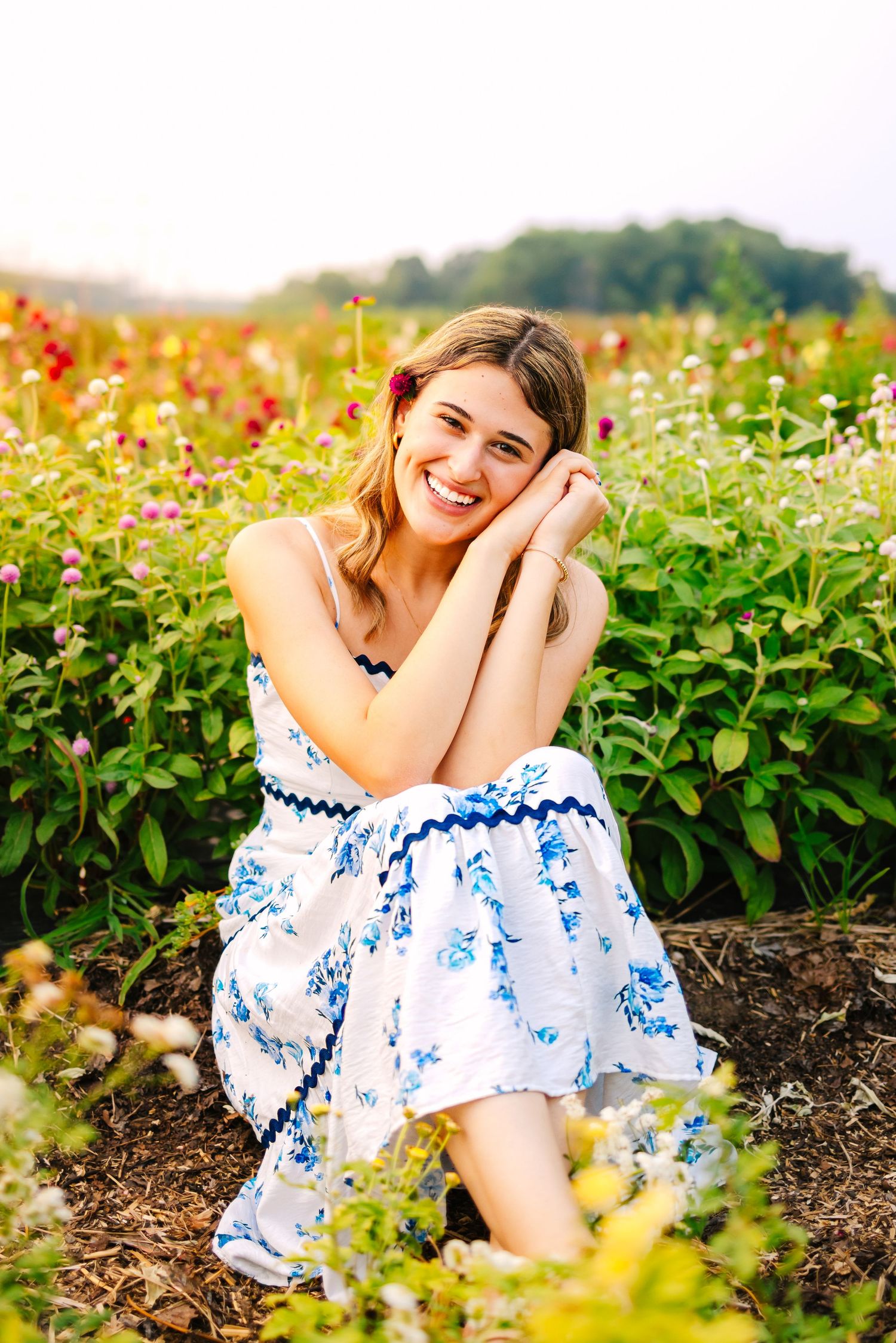 Joyful young woman in white floral dress sitting and laughing among vibrant blooming flowers in garden field.