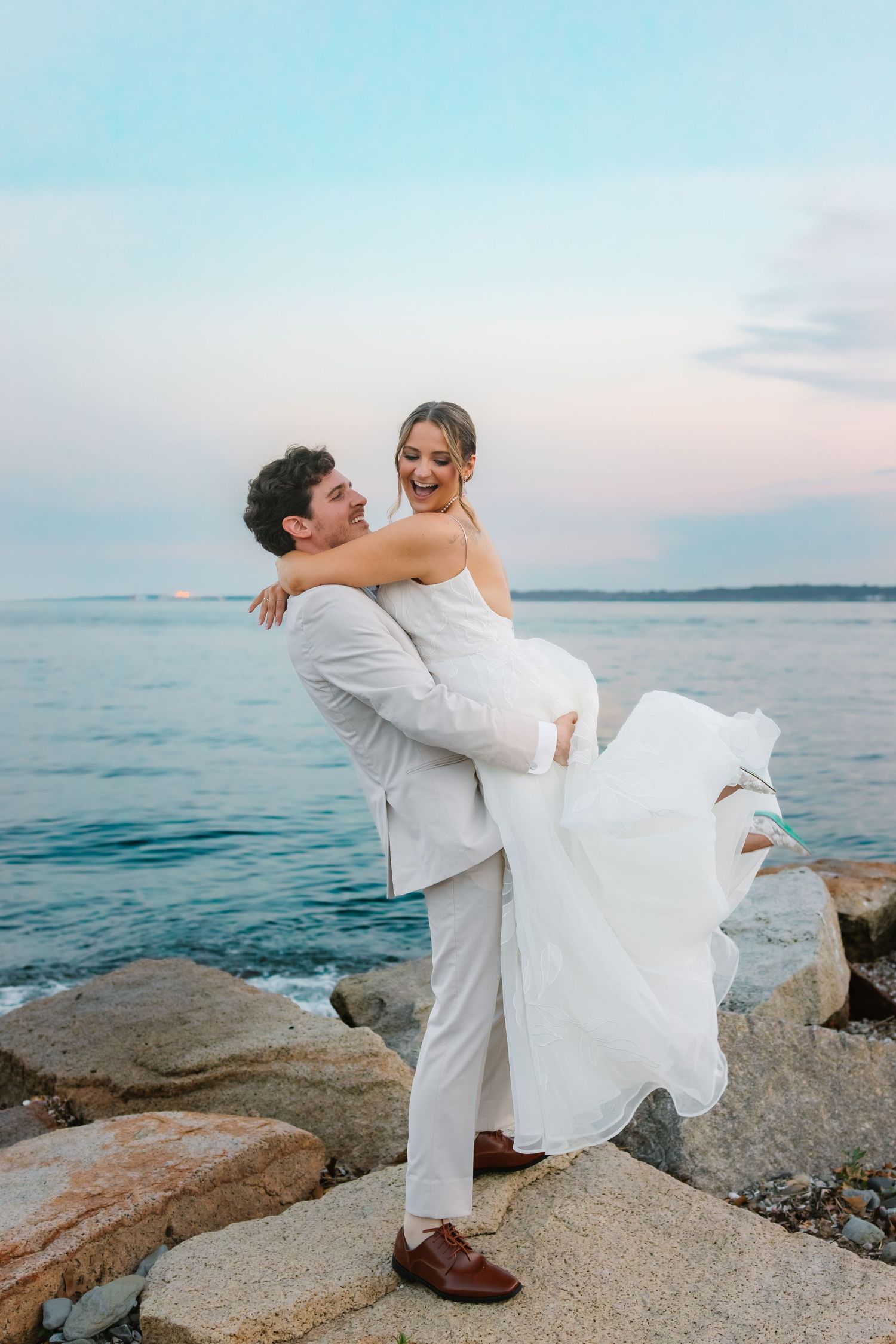 Groom lifting laughing bride in white wedding dress on rocky oceanside at dusk.