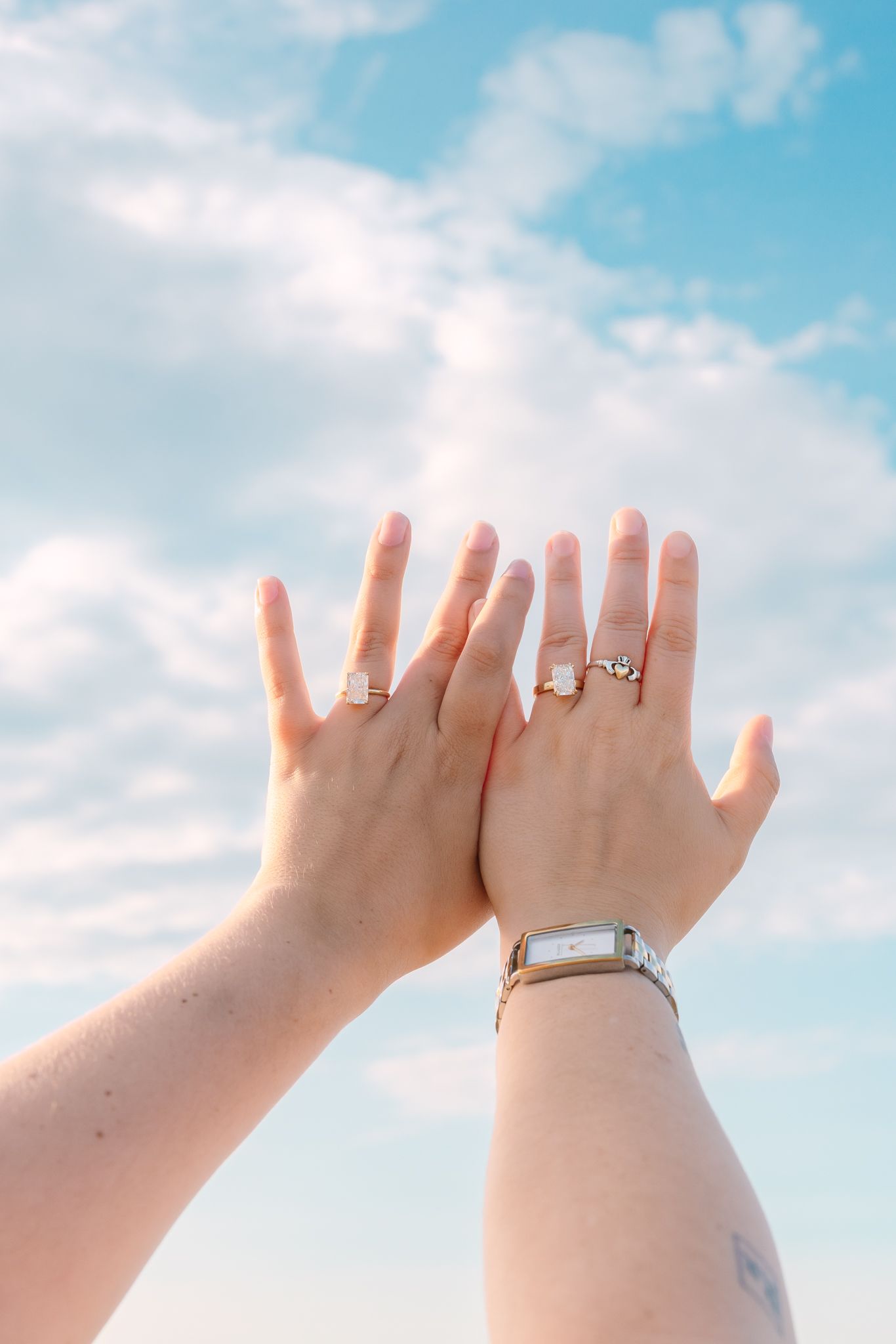 Two hands raised against blue sky displaying engagement rings after a same-sex beach proposal.