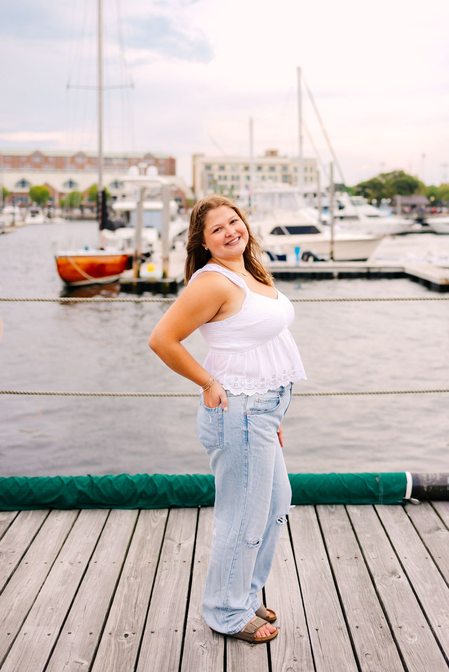 Young woman in white top and jeans smiling on a marina dock with sailboats in the background.