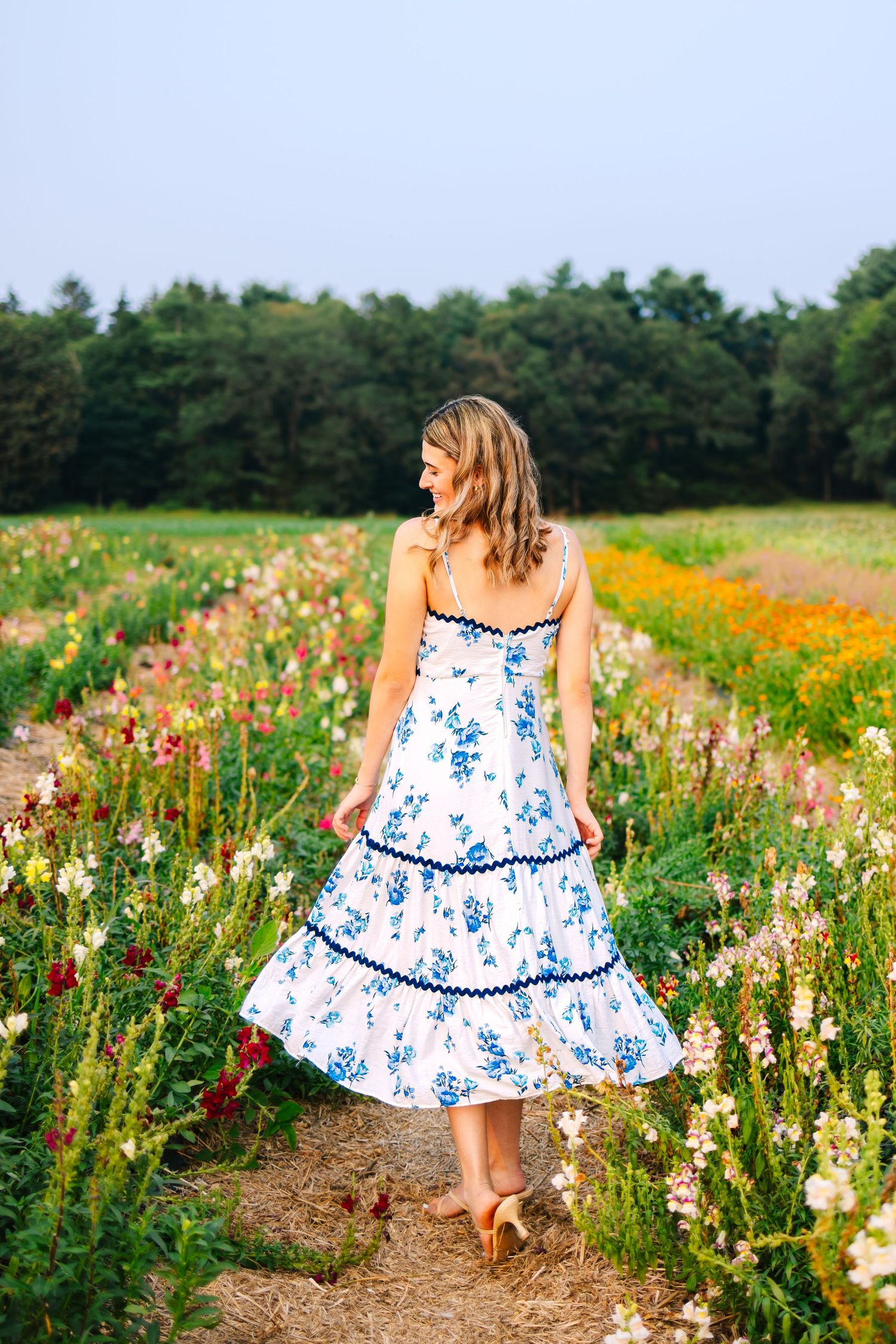 Back view of woman in white blue floral tiered midi dress standing in vibrant wildflower farm field.