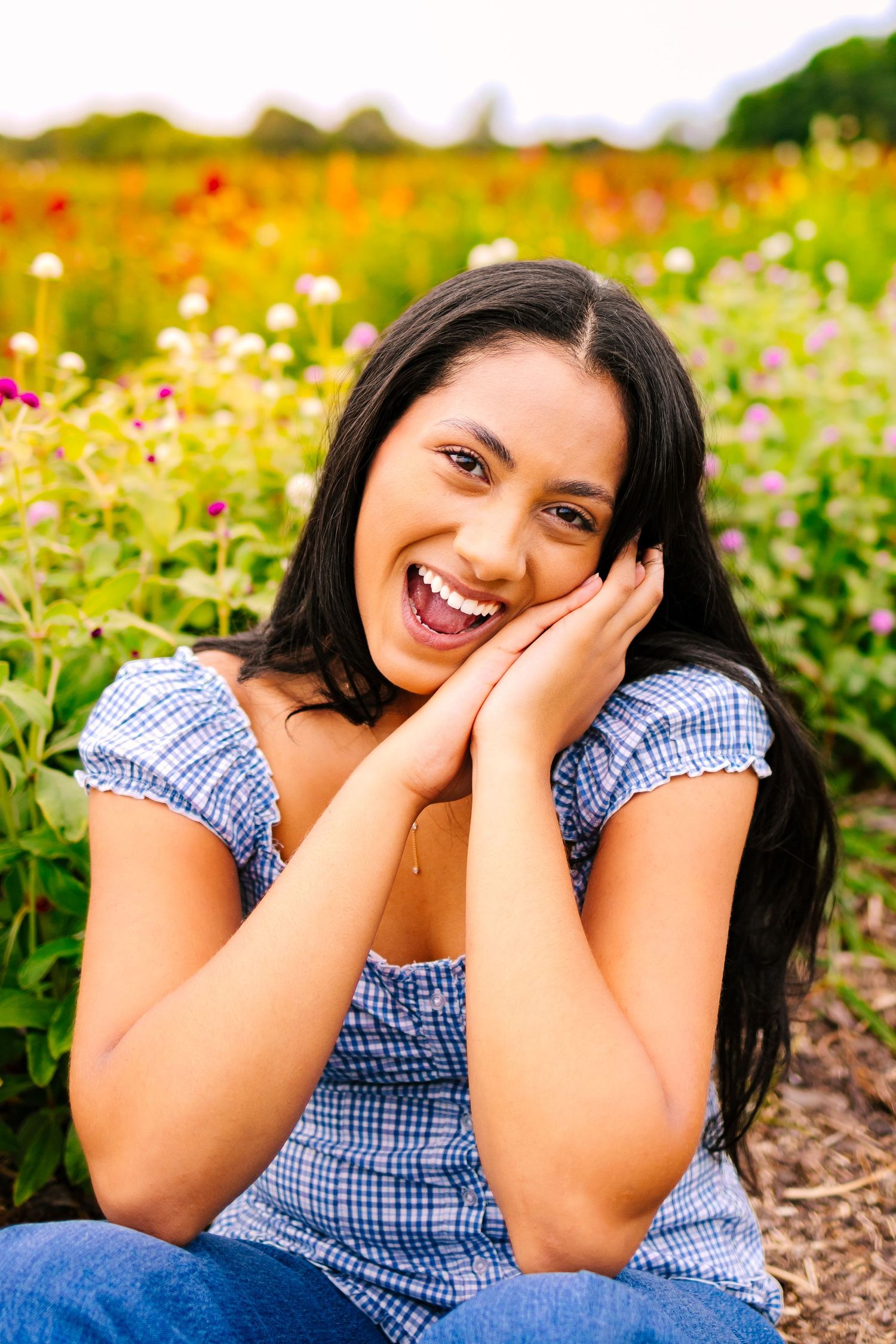 Joyful young woman in blue plaid top sitting with hands on cheeks in vibrant colorful flower field.