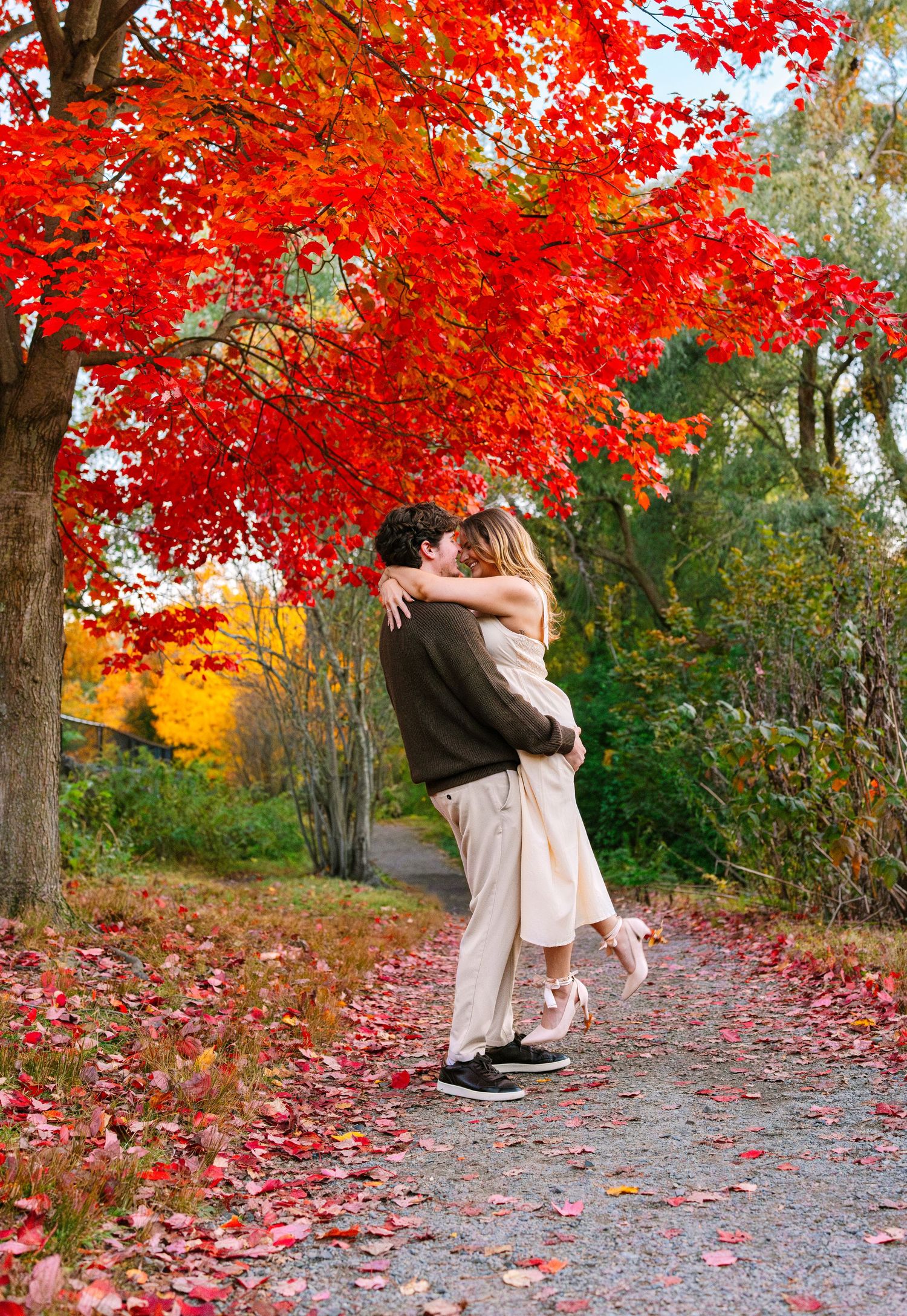 Two people embrace on a leaf-covered path surrounded by vibrant red autumn maple trees and golden fall foliage.