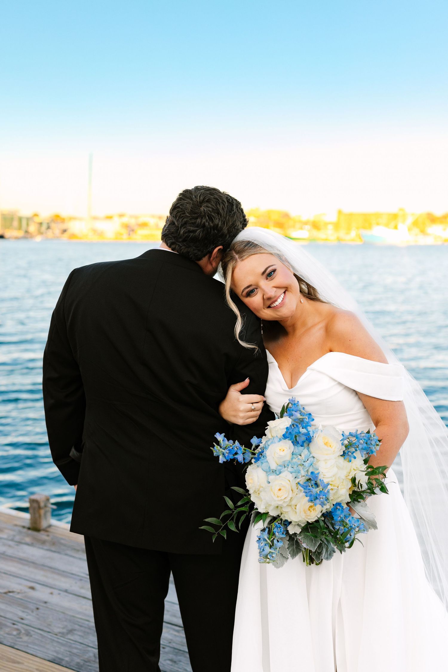 A couple shares a romantic moment on a dock with blue and white floral bouquet at sunset.