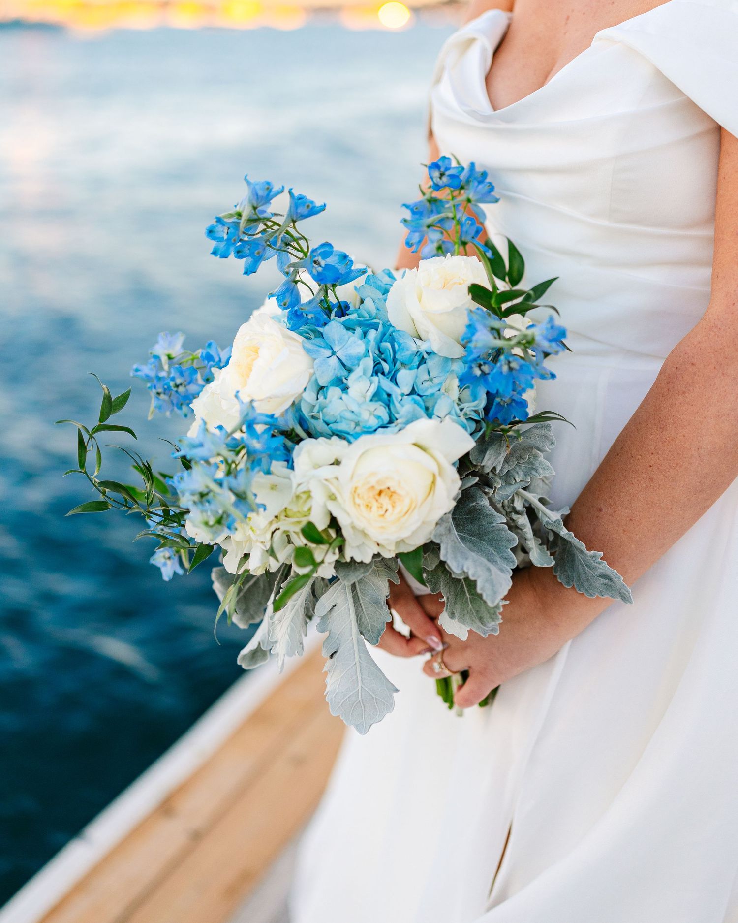 A bridal bouquet featuring white roses and blue delphinium flowers against an ocean backdrop at The Regatta Place in Newport Rhode Island