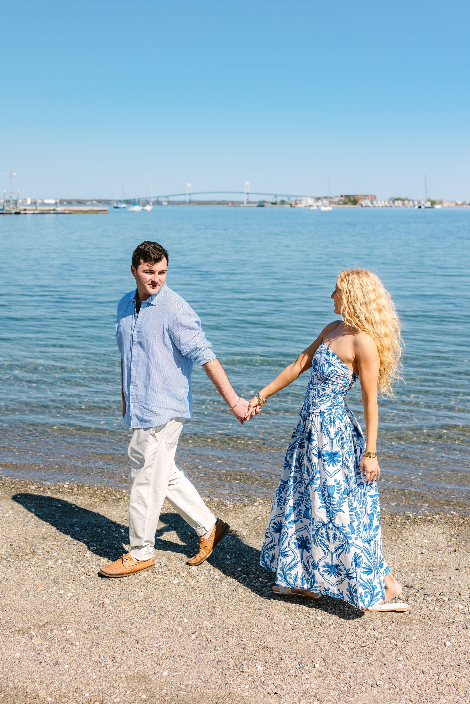 Happy couple holding hands and walking along sunny beach shoreline with bridge visible in background.