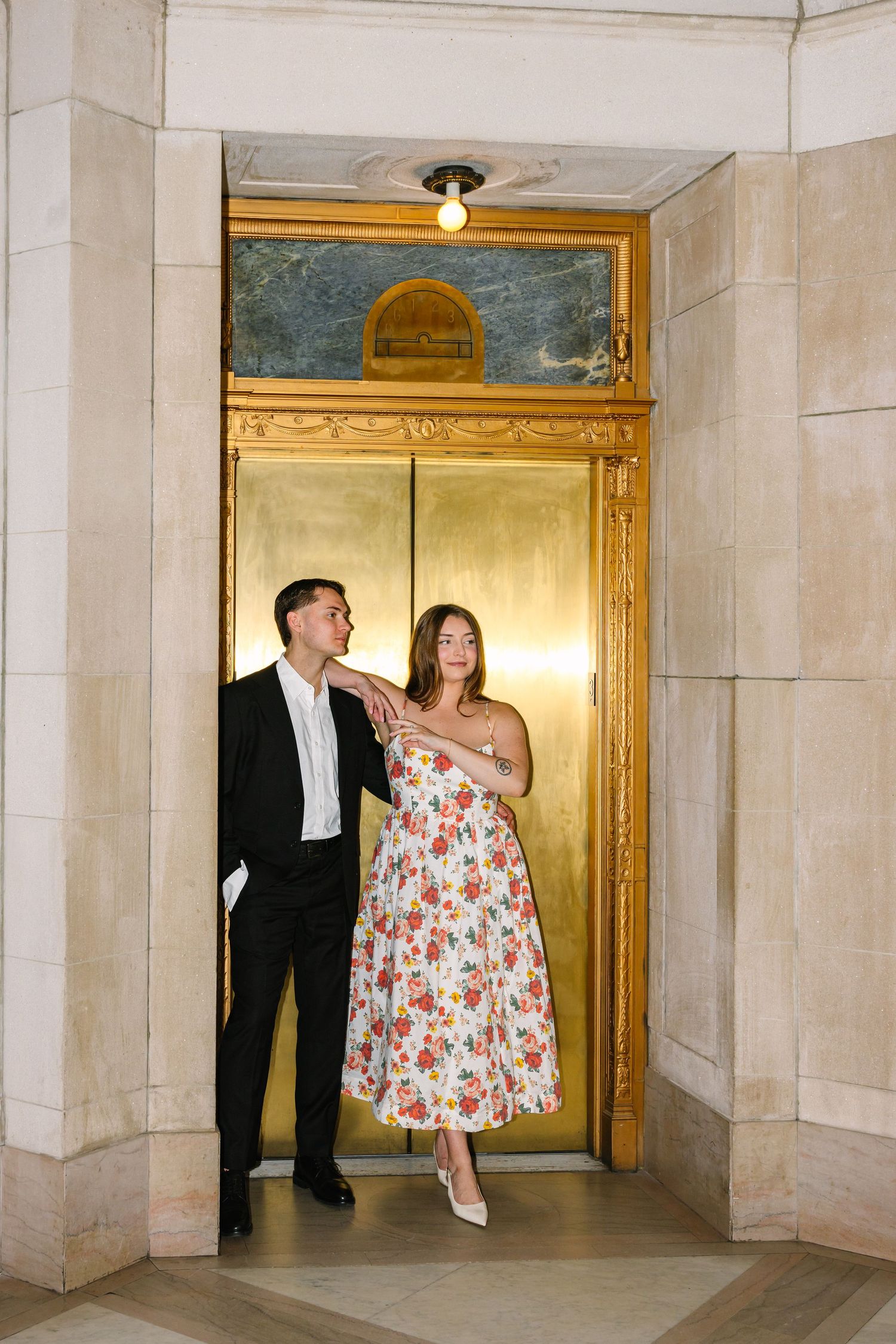 Couple posing together in front of stunning gold elevator doors in an elegant marble building.