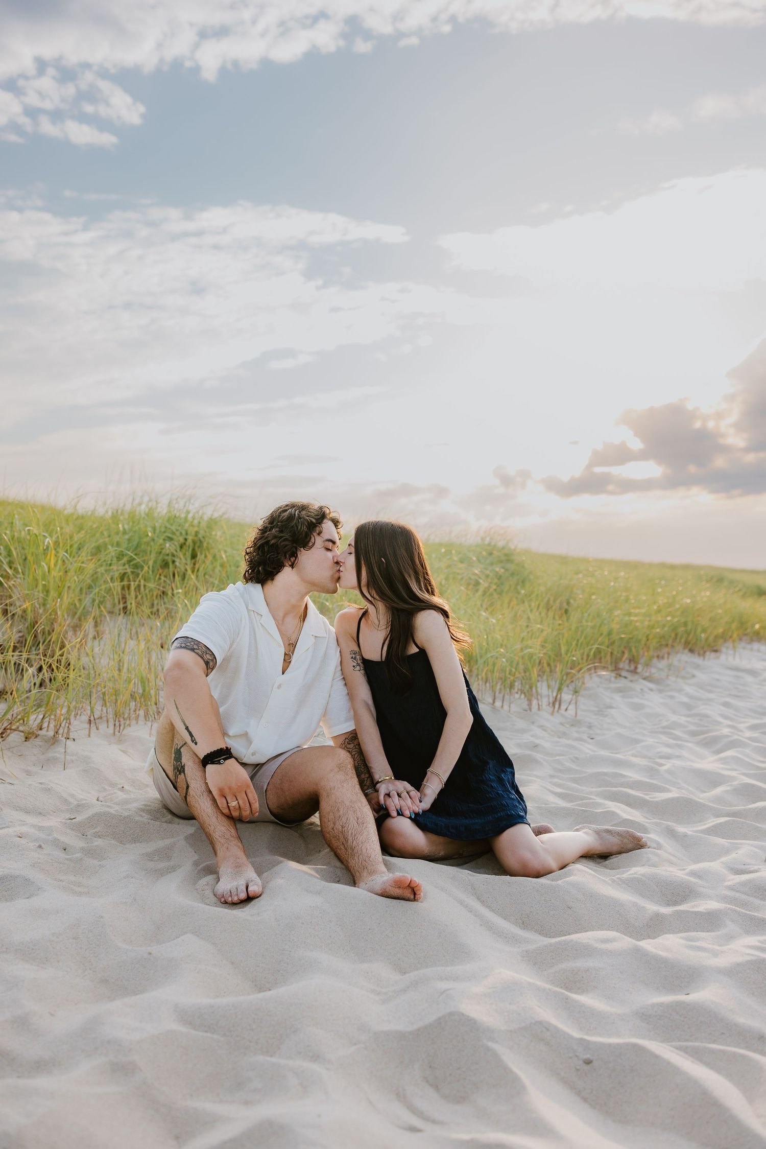 Couple sharing a tender kiss while sitting on sandy beach dunes with grass and cloudy sky.
