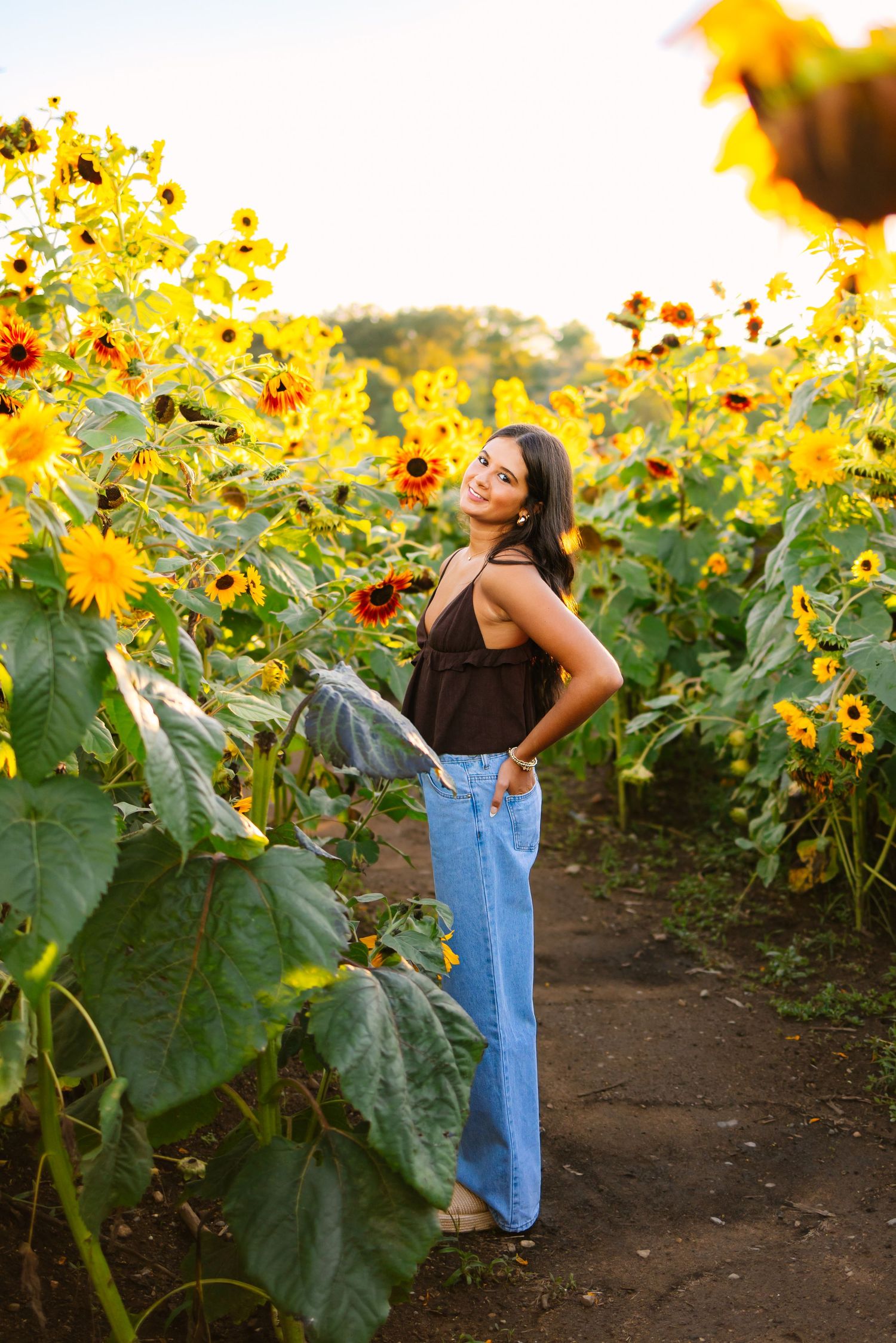 Young woman in black top and jeans poses among vibrant yellow sunflowers in sunny field.