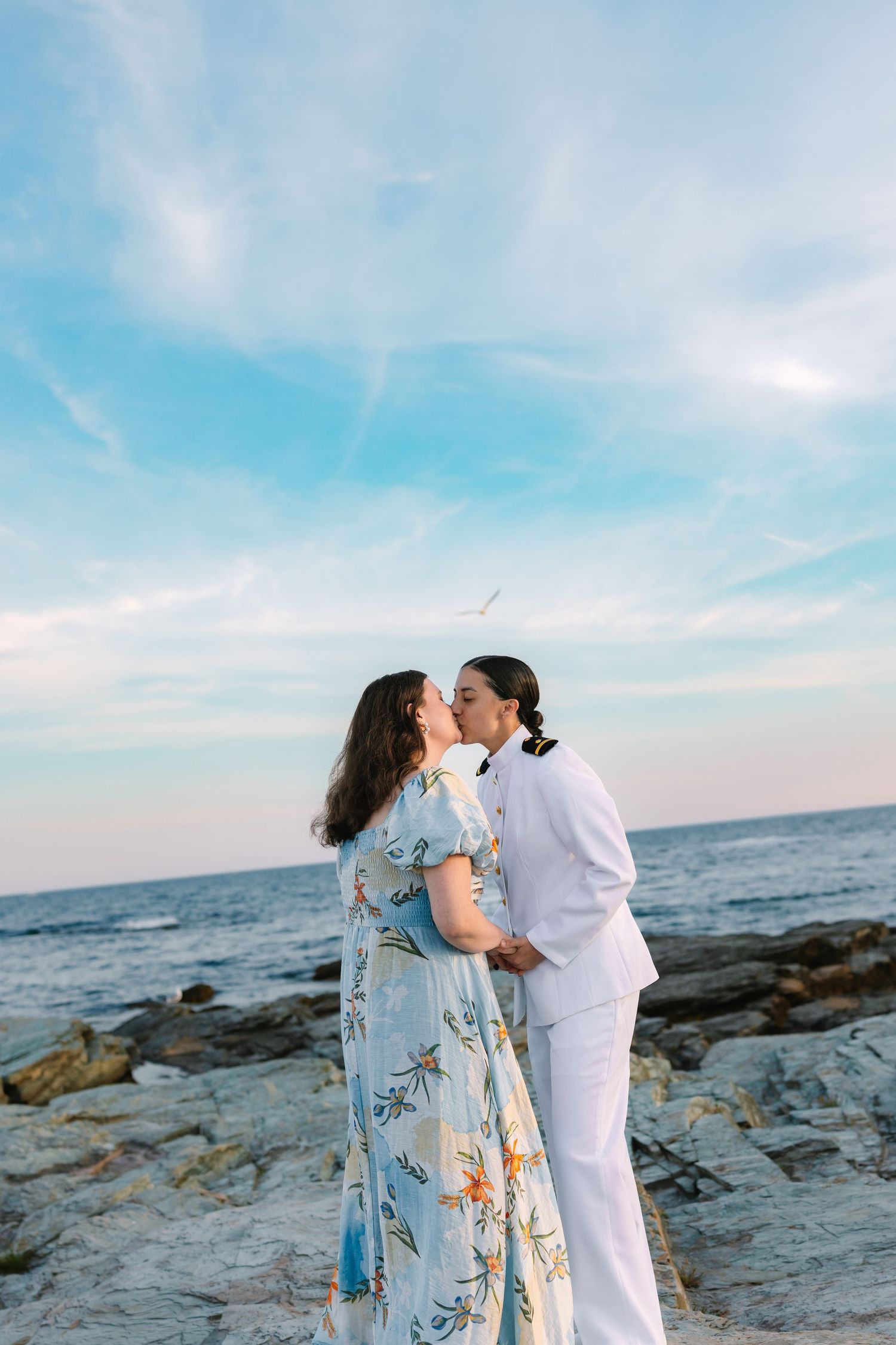 A couple shares a romantic kiss on a rocky beach with blue sky and ocean backdrop.