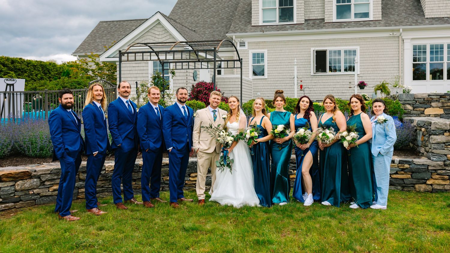 Wedding party in blue suits and emerald green dresses poses together on a lawn in front of white house.