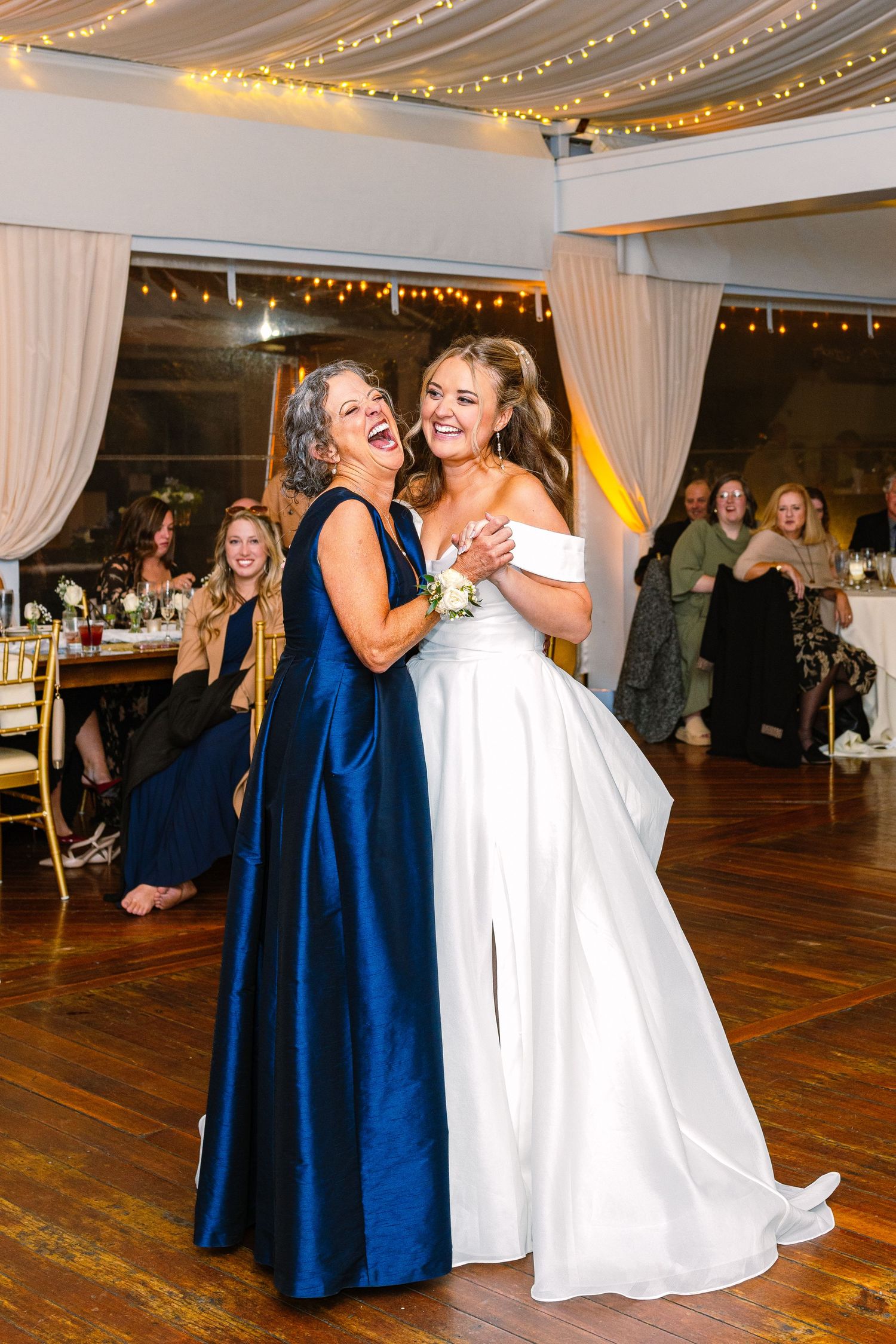 Two people share an emotional dance moment at a wedding reception with string lights and navy bridesmaids dresses visible.