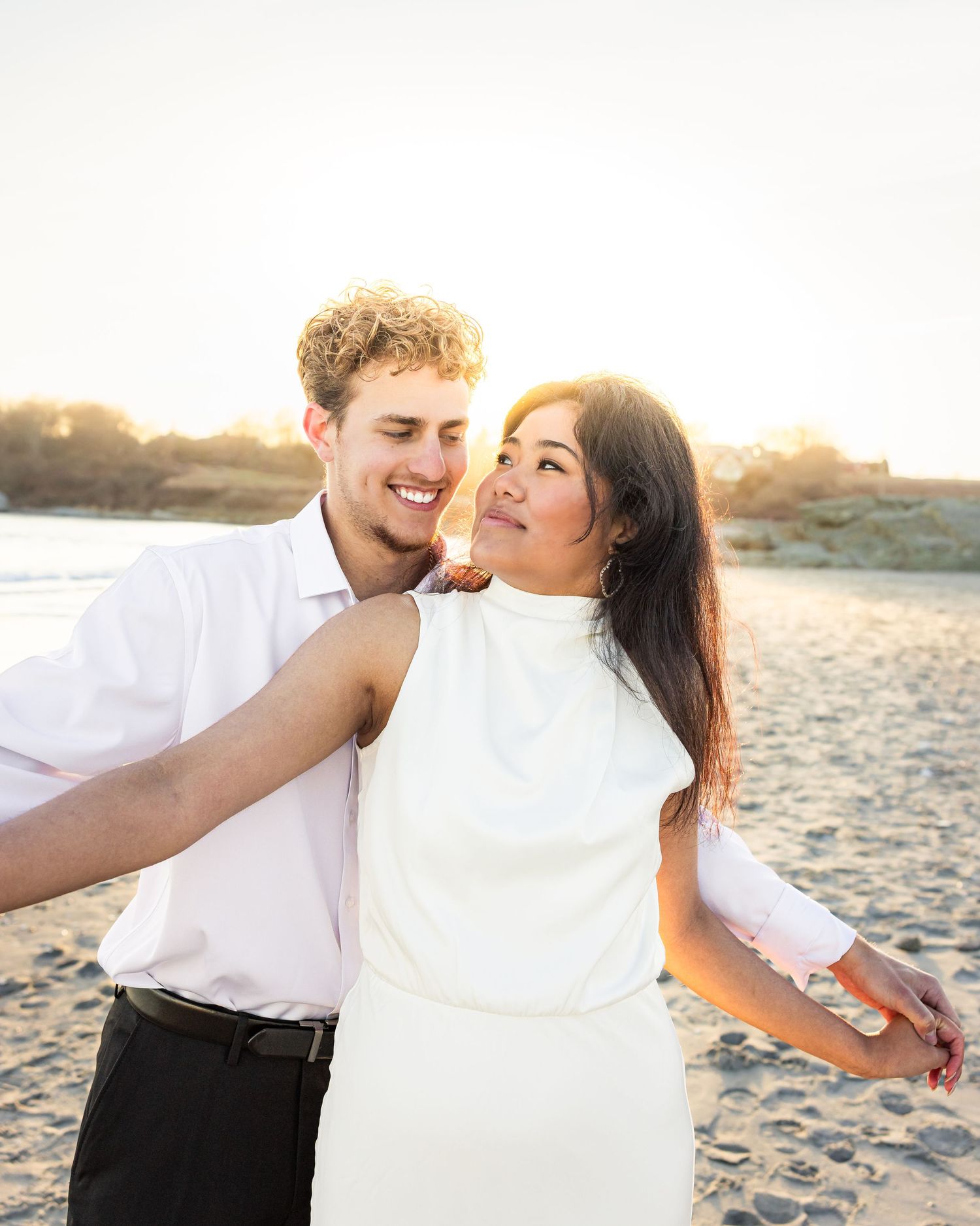 A couple shares a joyful moment together on the beach at sunset, dressed in white outfits.