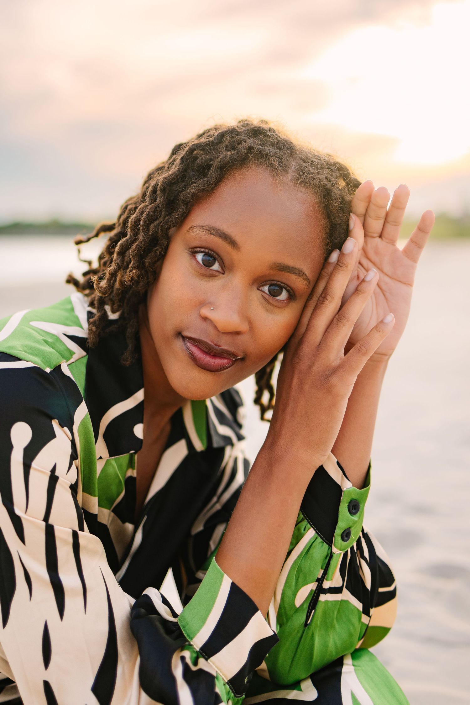 Beautiful Black woman in tropical print shirt posing thoughtfully at beach during colorful sunset.