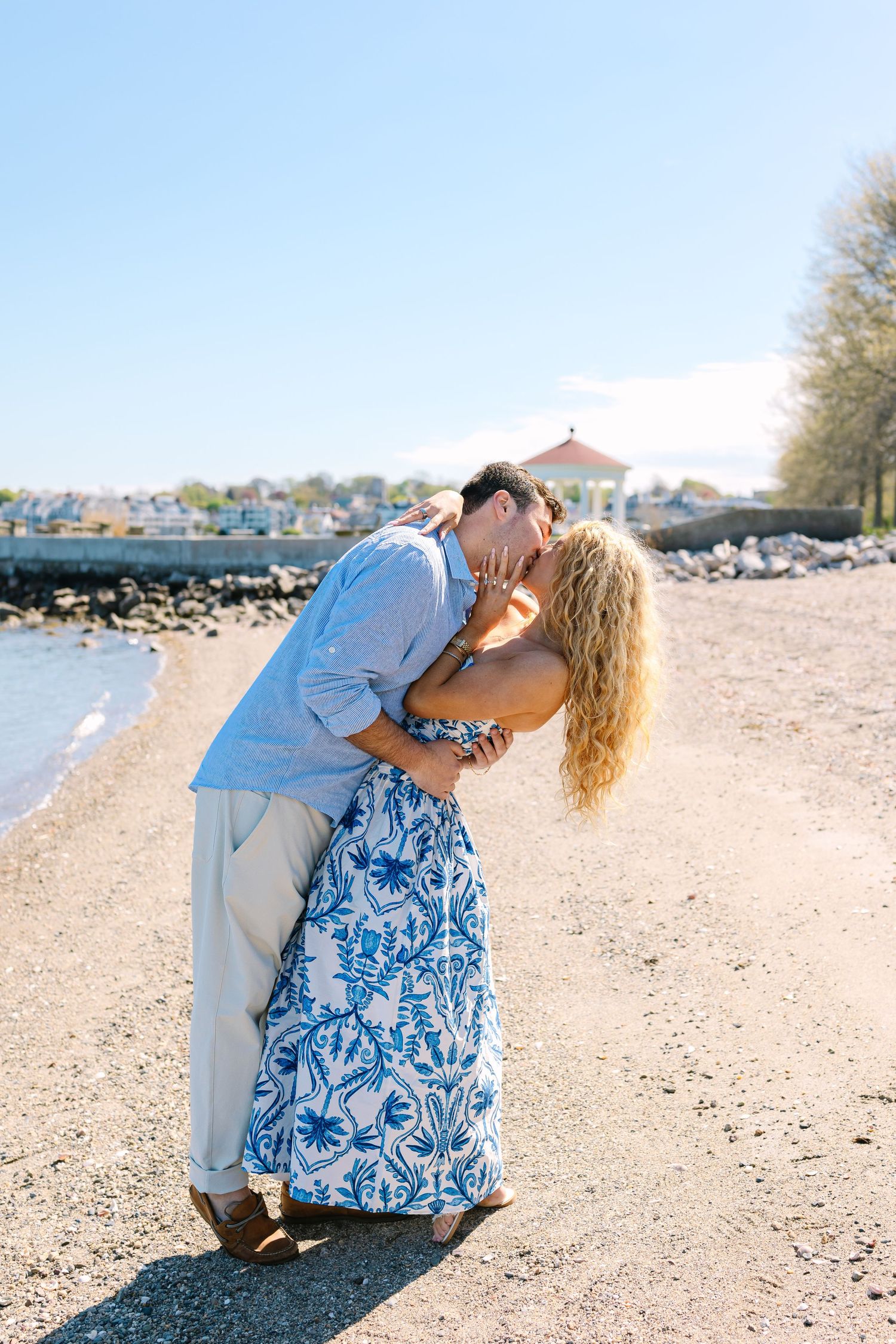 Couple sharing a passionate dip kiss on a sunny beach with a red gazebo visible in the background.