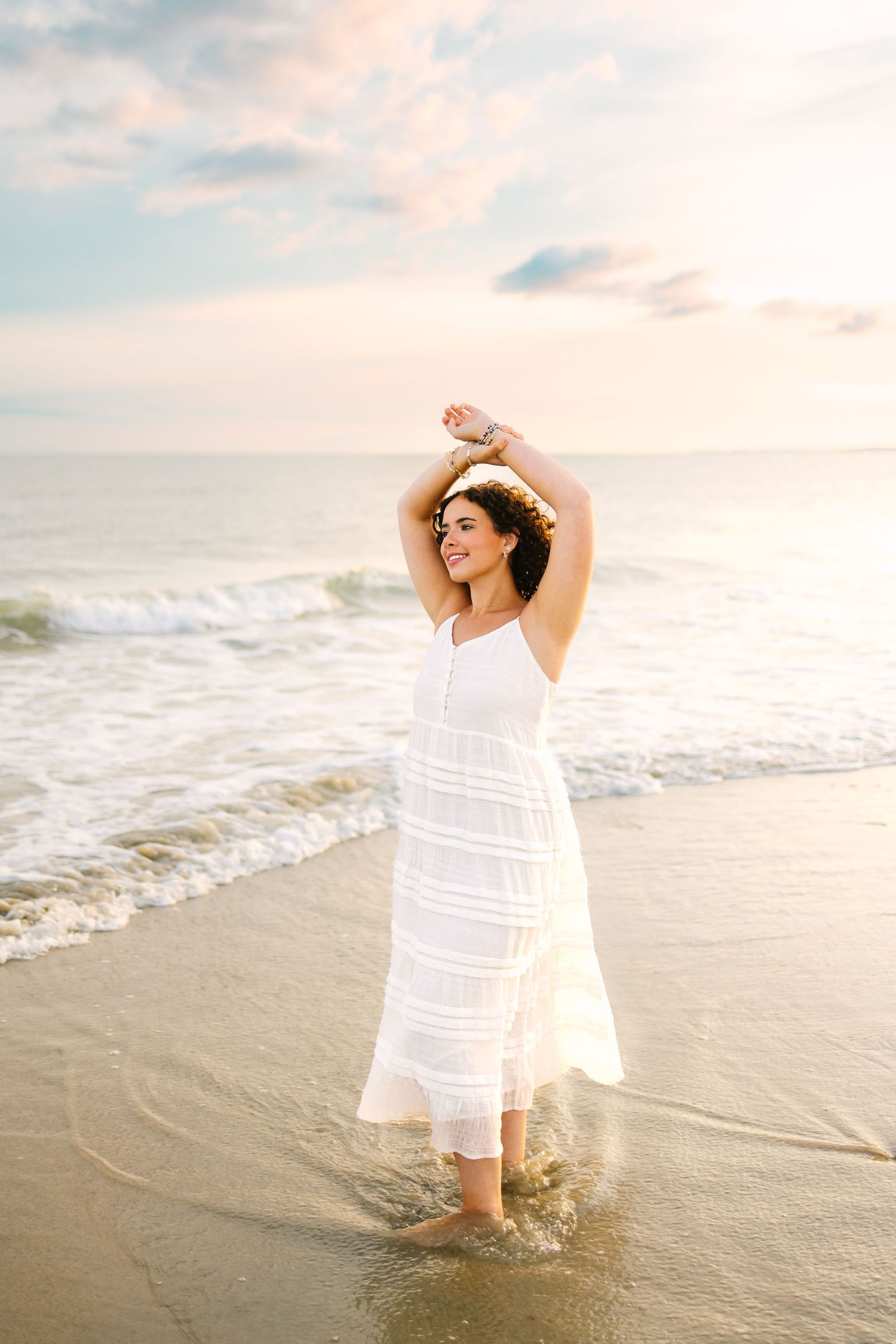 Woman in white dress raising arms joyfully while standing in ocean waves at sunset.
