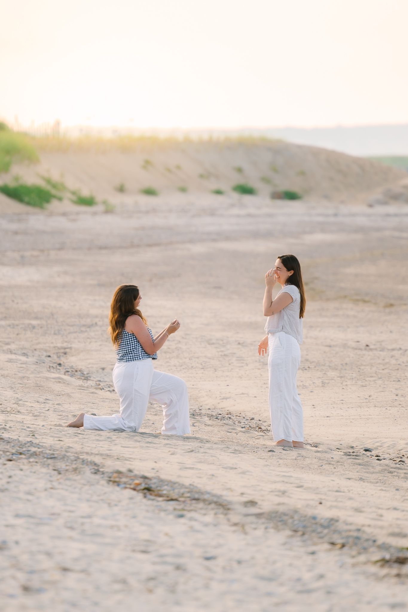 Emotional same-sex marriage proposal on sandy beach at golden hour, woman on one knee proposing.
