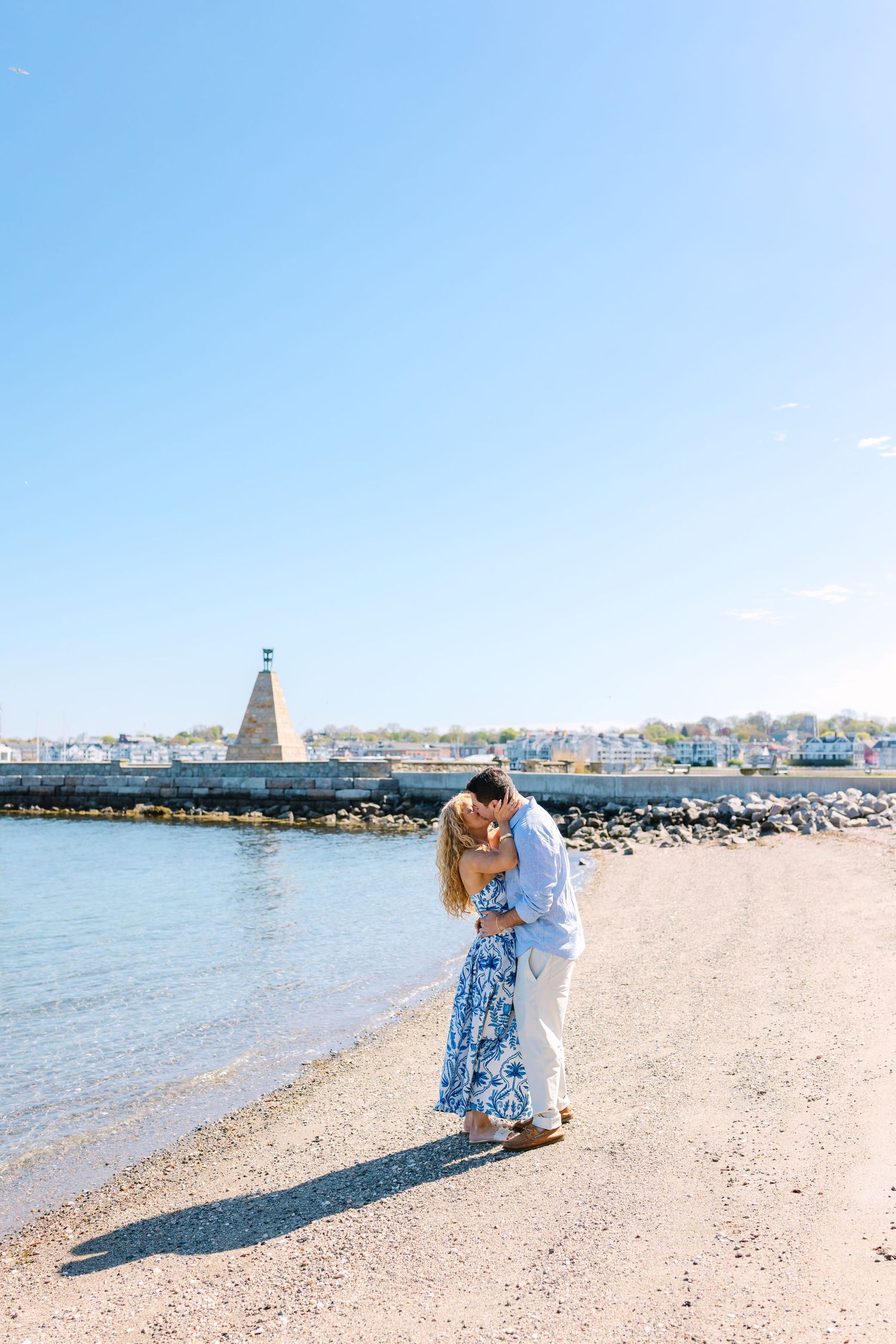 Couple sharing a romantic dip kiss on a sunny beach with a stone monument and harbor in the background.