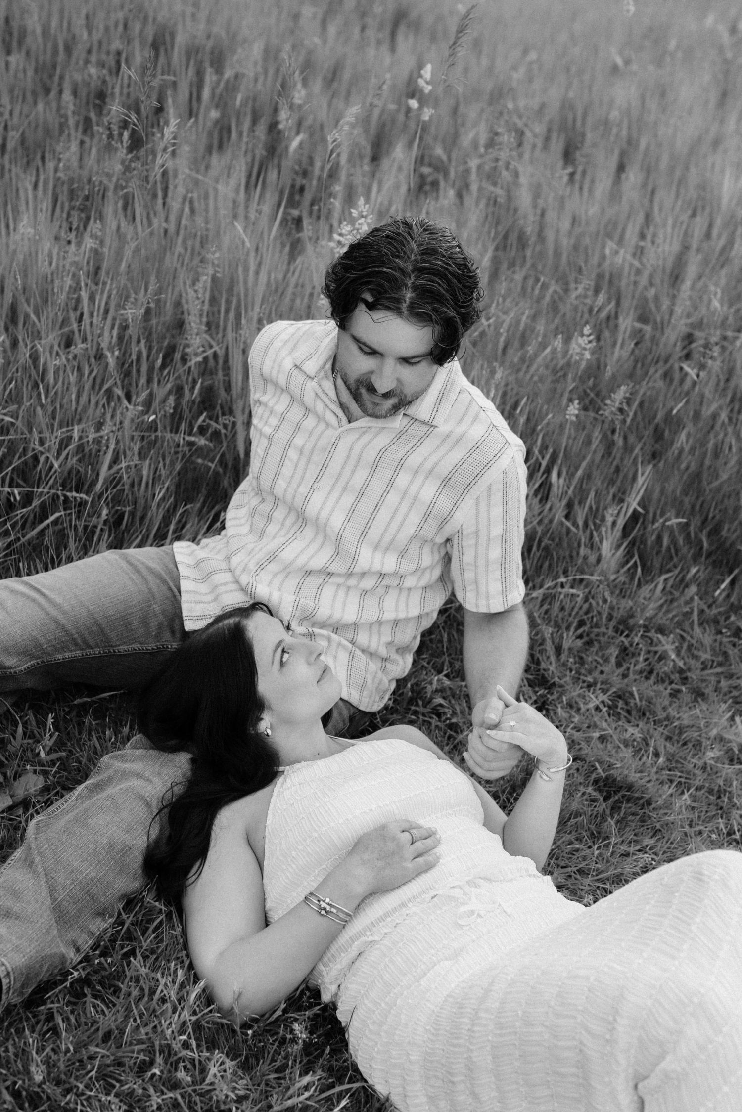 Black and white photo of a couple in a meadow, woman lying in grass smiling up at her partner.