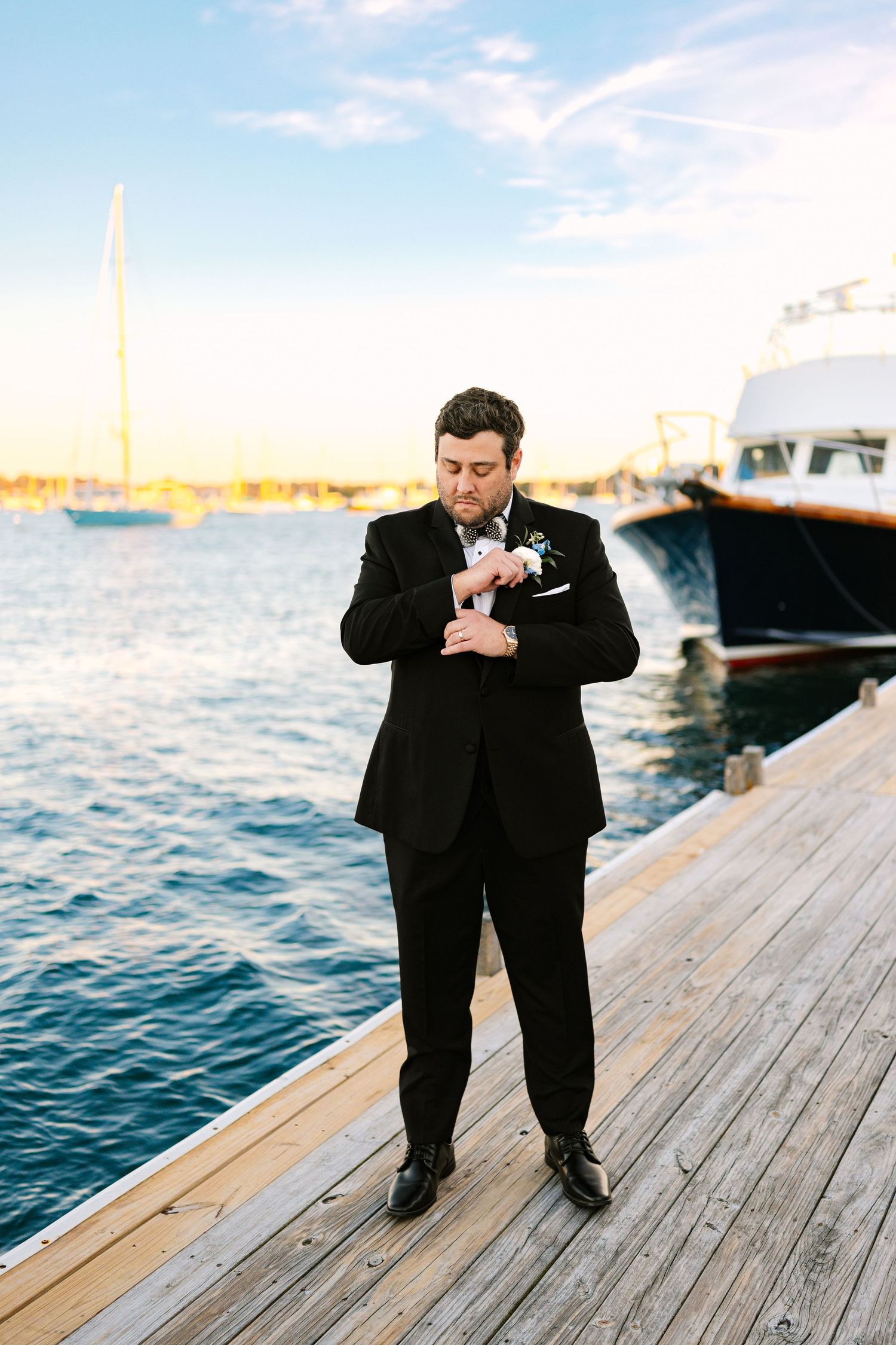 Groom in formal black attire fixing his suit on a wooden dock by the ocean at sunset at The Regatta Place in Newport Rhode Island