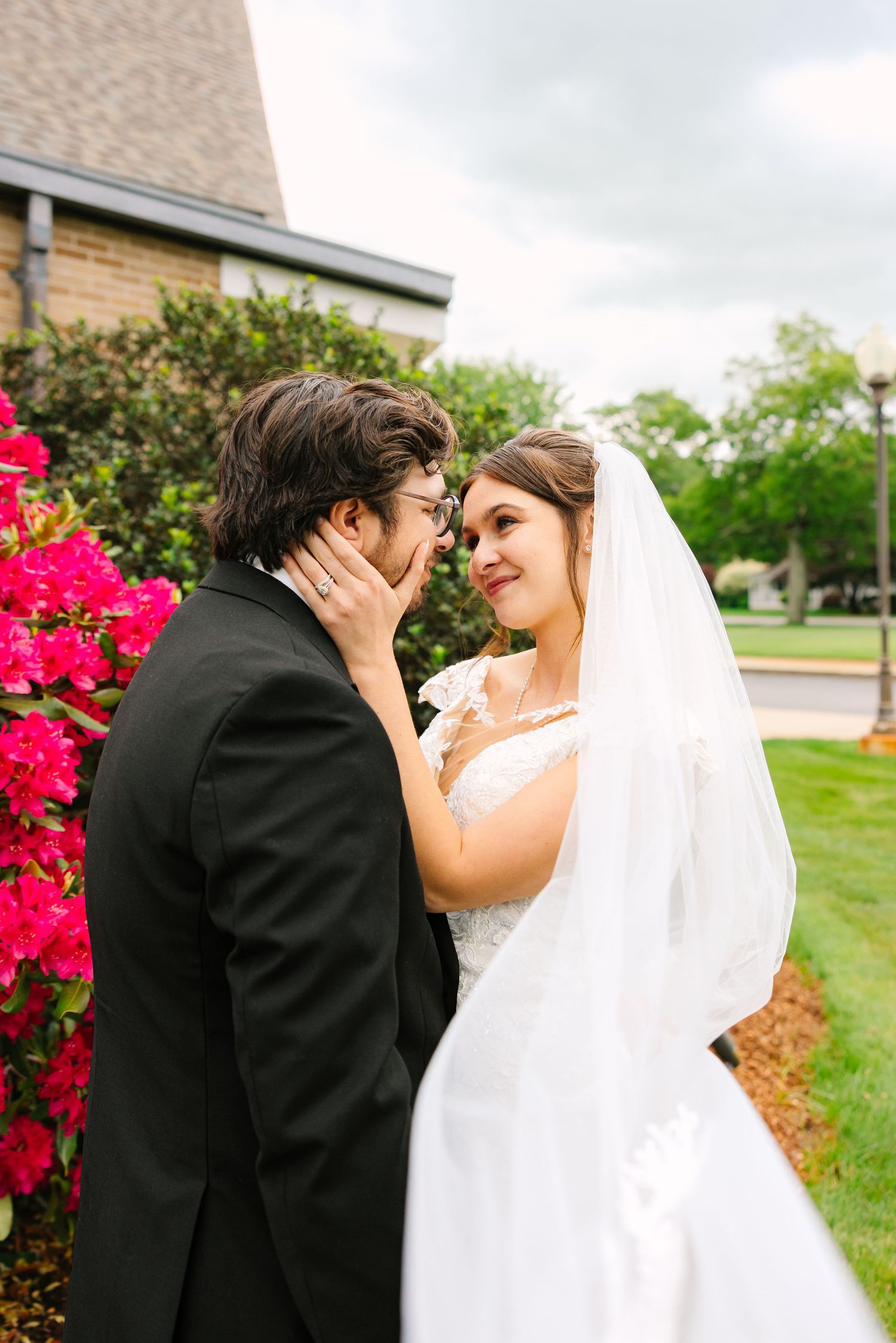 Bride and groom sharing tender almost-kiss moment near vibrant pink azalea bushes outside church.