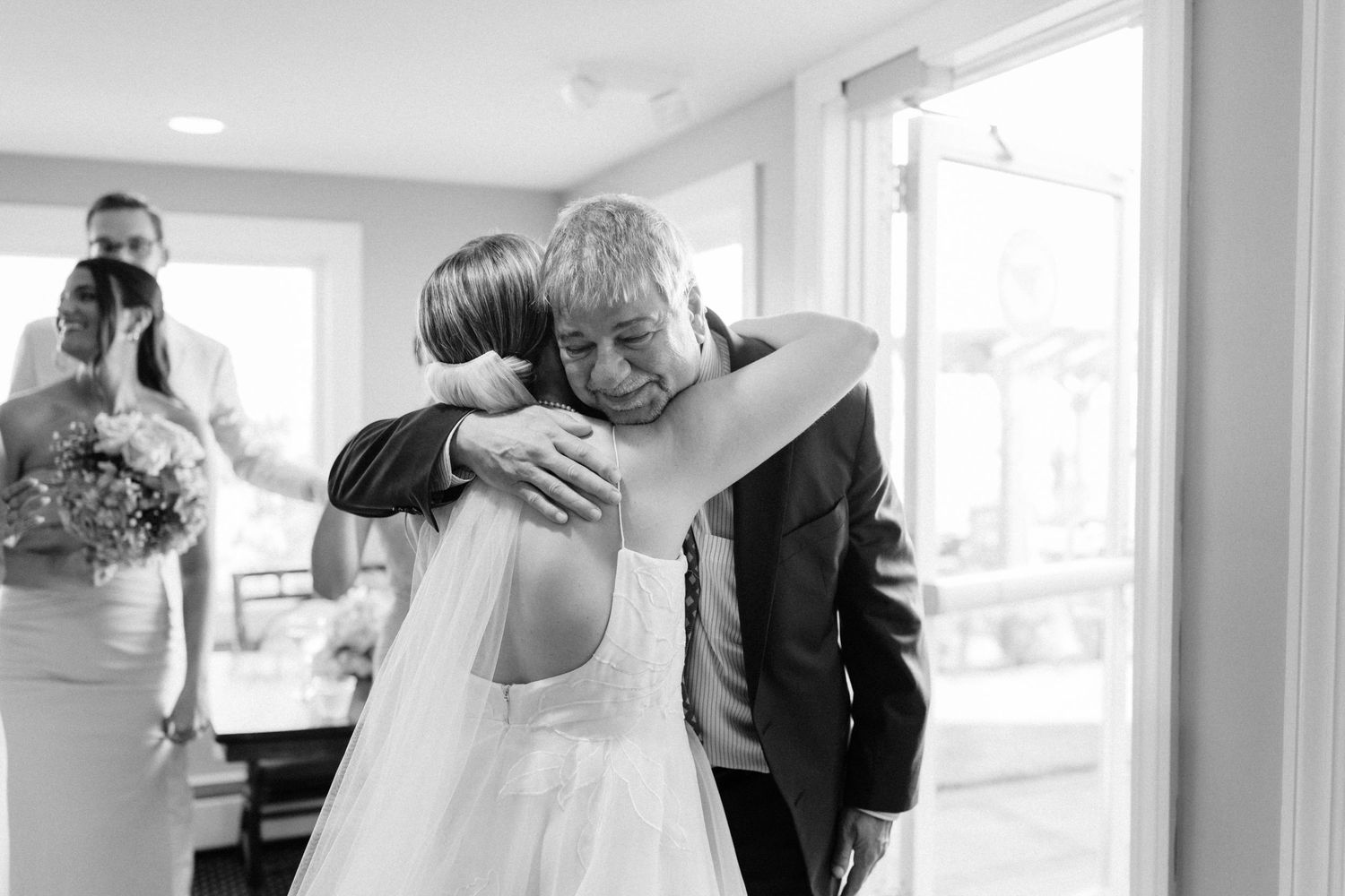 Black and white photo of bride in wedding gown sharing a warm embrace with an older gentleman before the ceremony.