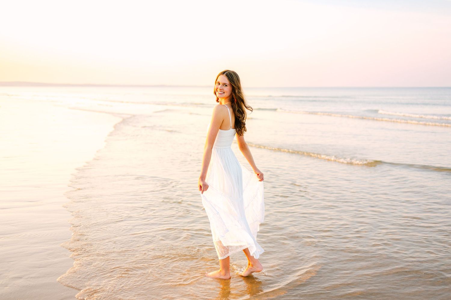 A happy young woman in a white strapless dress walks along a serene beach at golden sunset.