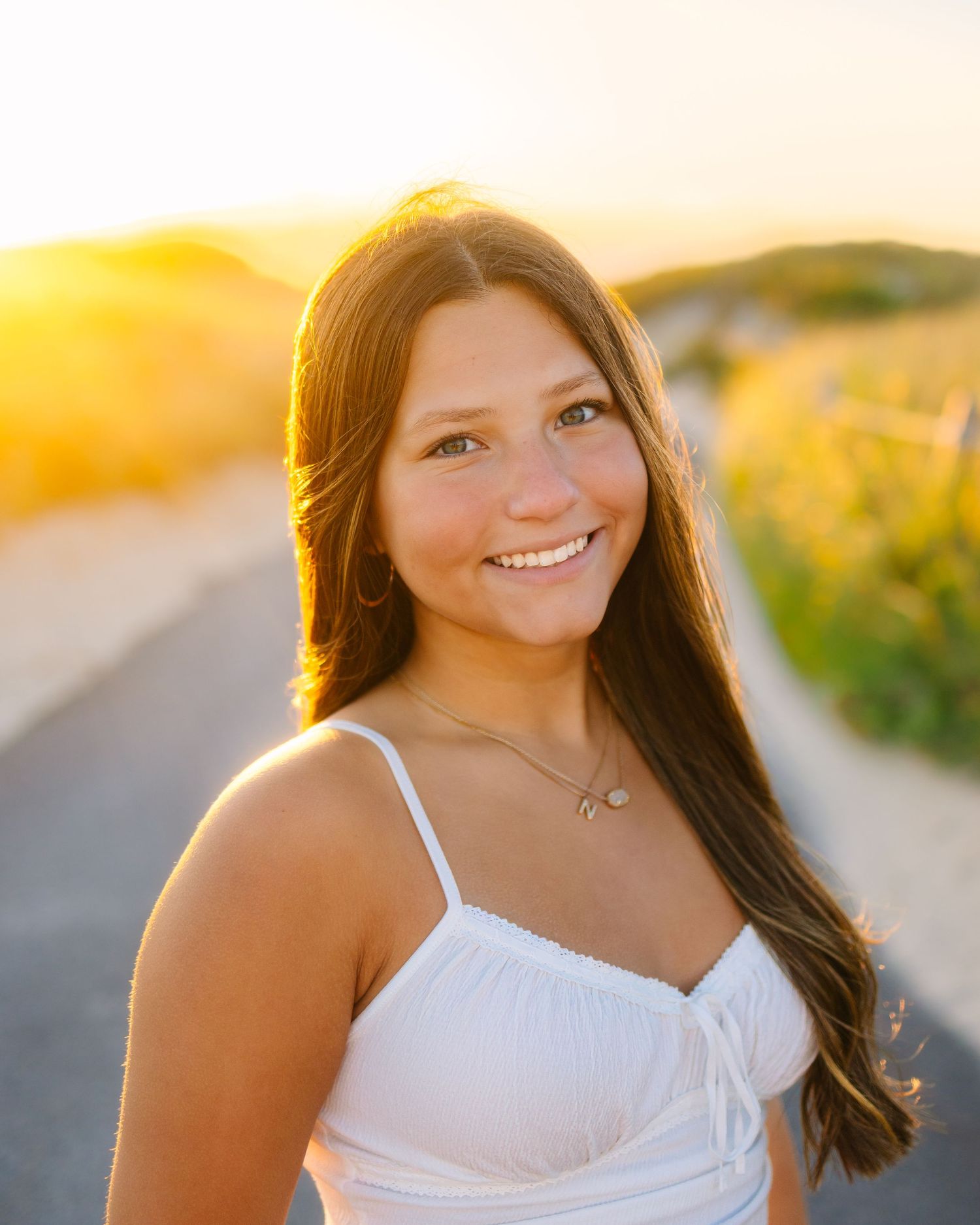 Smiling teen girl in white sundress poses on sandy path during warm golden hour senior portrait.