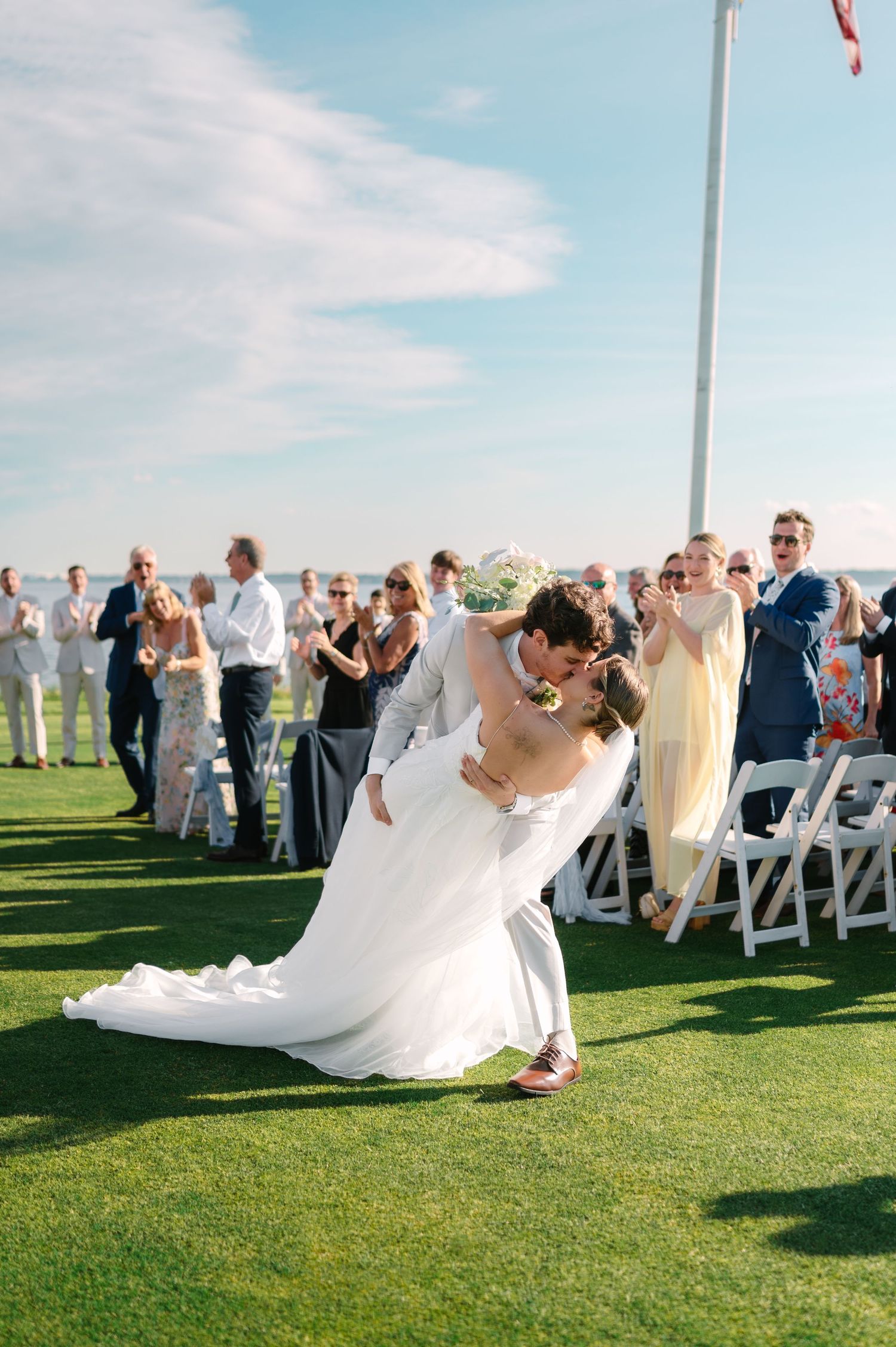 Groom dips bride in a stunning white gown as wedding guests cheer during a joyful outdoor ceremony recessional on green lawn.
