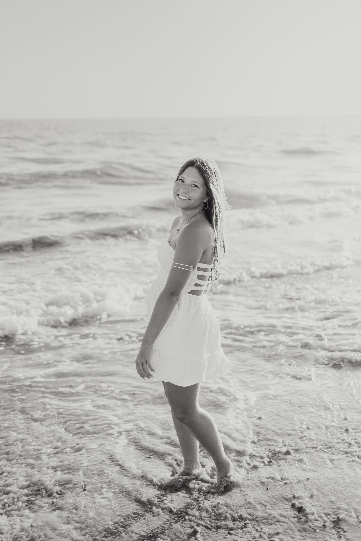 Black and white photo of smiling young woman in white dress standing in ocean waves on beach.