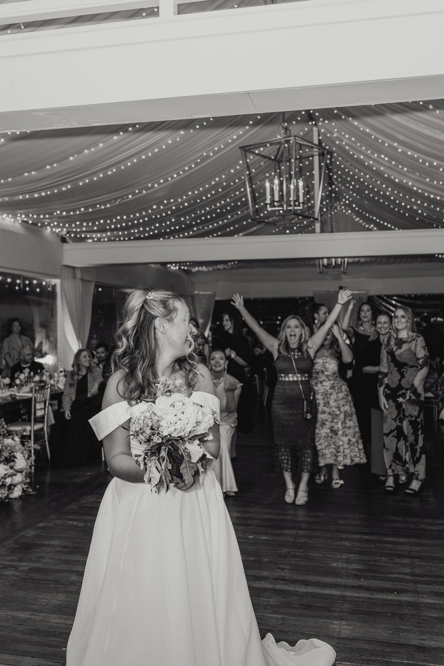Black and white wedding reception photo showing dancing guests under string lights while bride holds bouquet on dance floor.