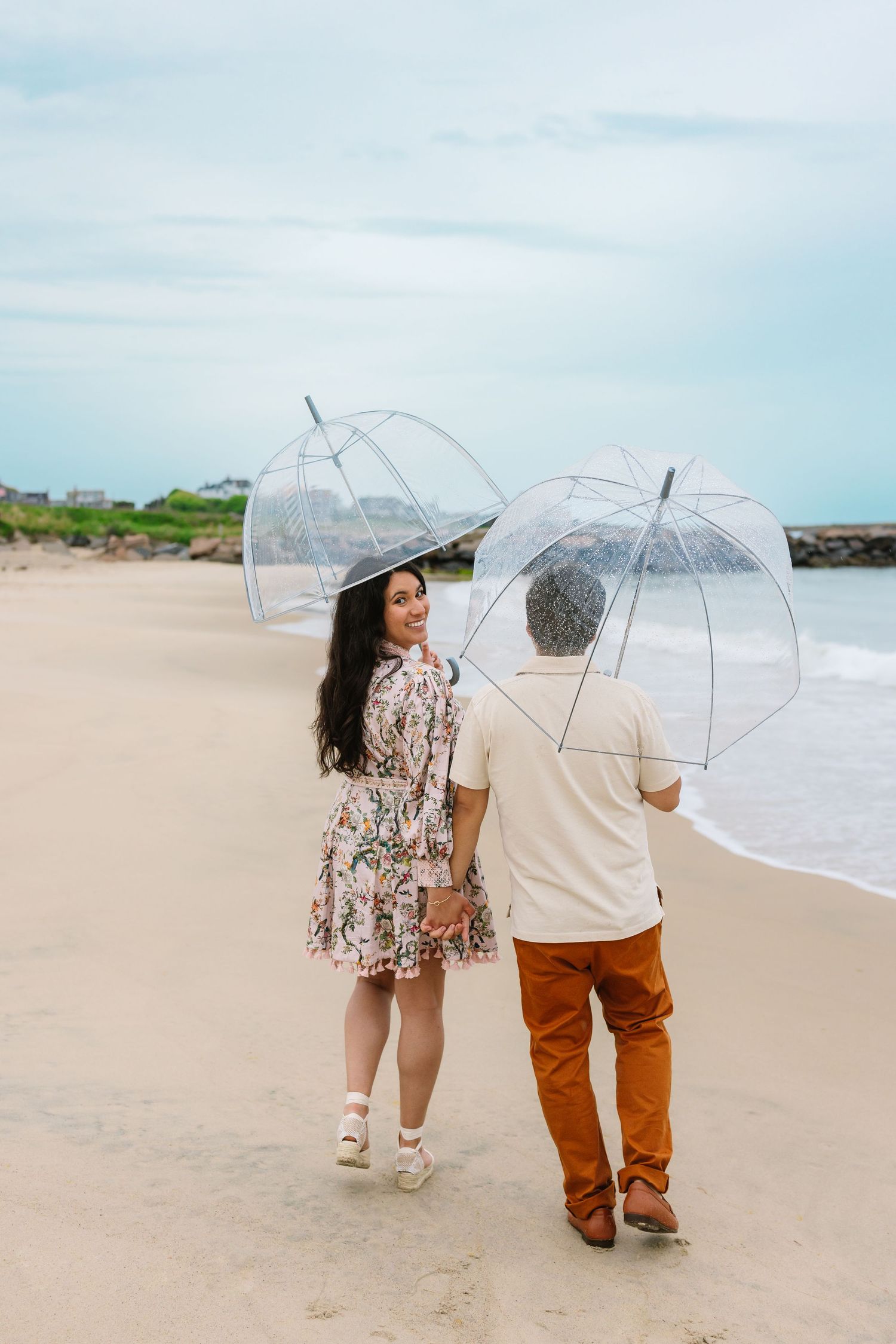 Couple strolling on a beach holding clear bubble umbrellas with rocky coastline ahead.
