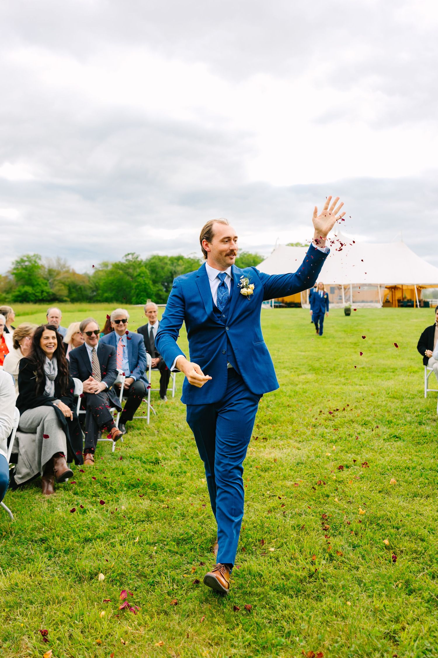 A groom in a bright blue suit celebrates joyfully while walking down a grassy aisle at an outdoor wedding ceremony.
