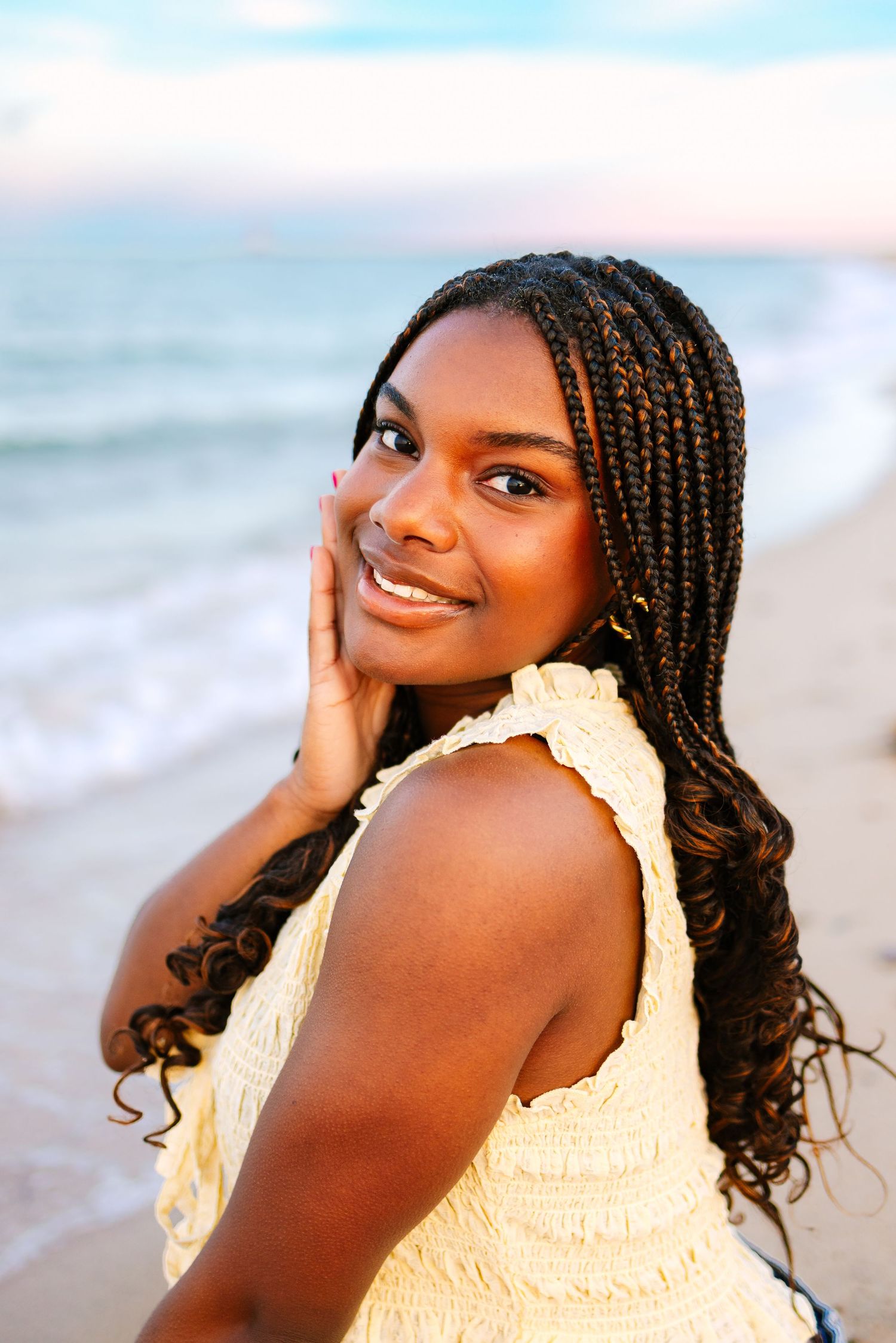 Young woman with braids in cream top smiles over shoulder on sandy beach with ocean waves behind her.